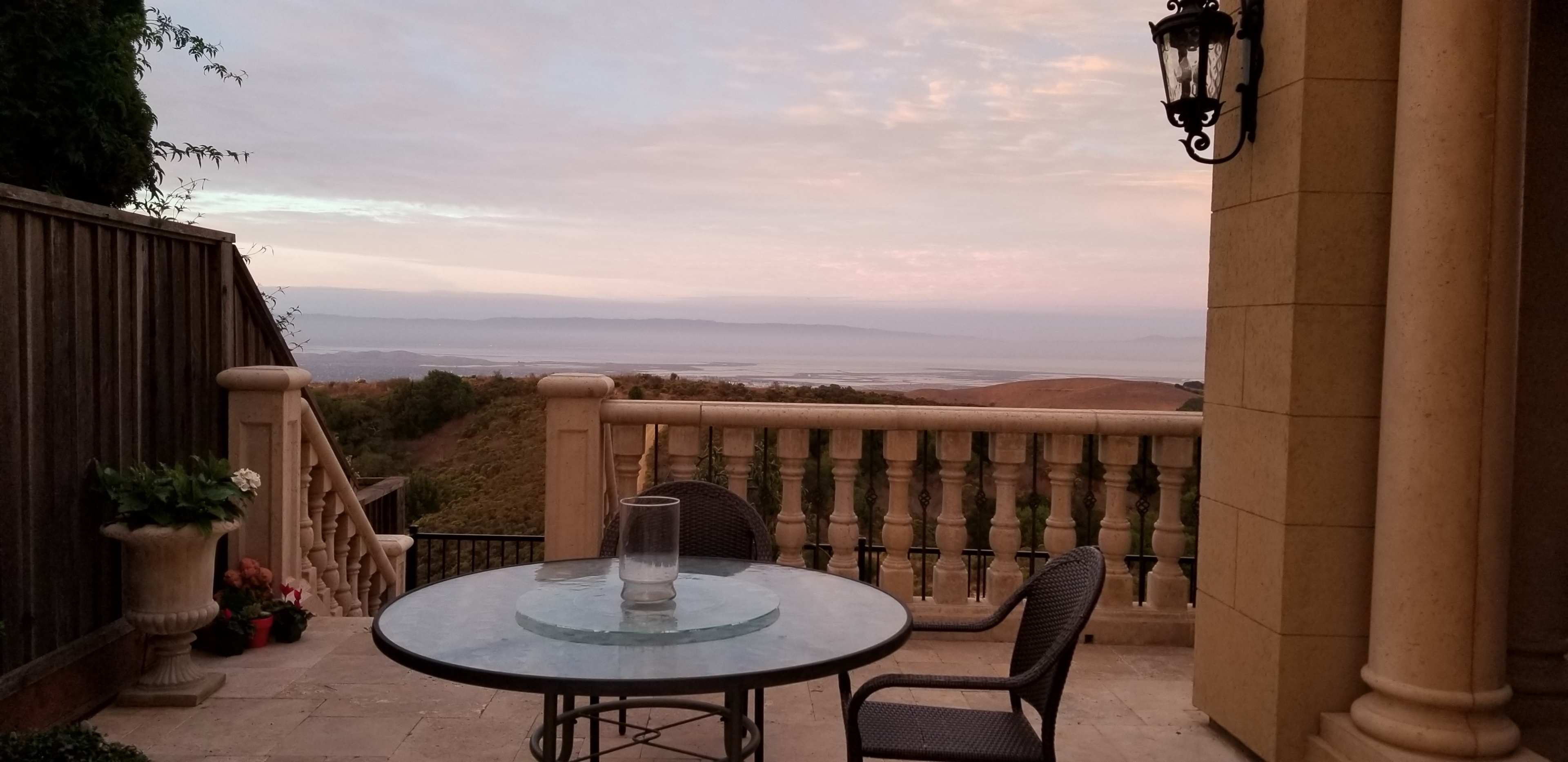 The image shows a balcony with a glass table and chairs overlooking a calm landscape and a body of water under a pastel sky.