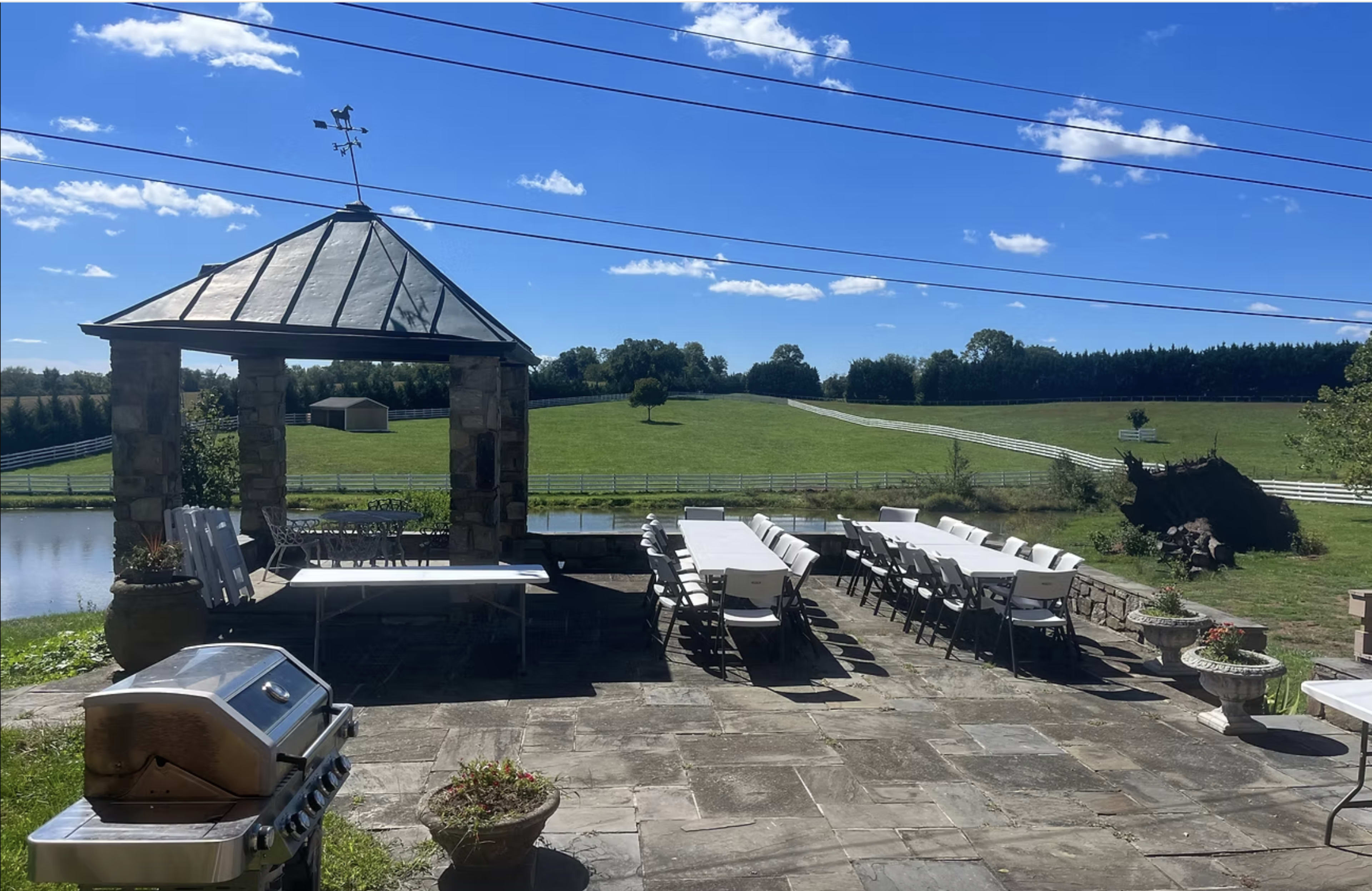The image shows a stone patio with a gazebo, several long tables arranged for an outdoor event, and a scenic view of green fields and a blue sky in the background.