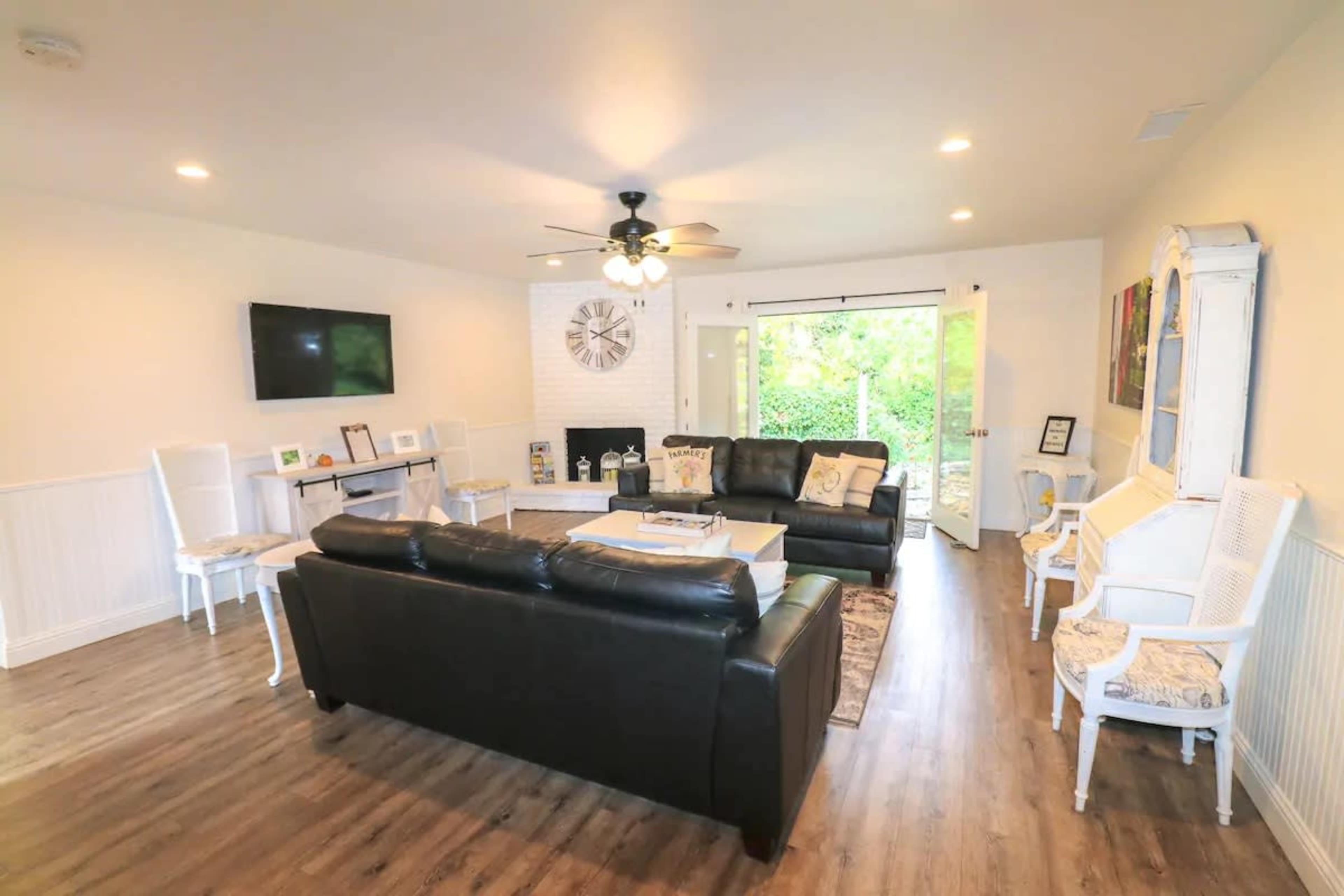 A spacious living room with a black leather sofa, a wall-mounted television, and large sliding doors leading to a green outdoor area.