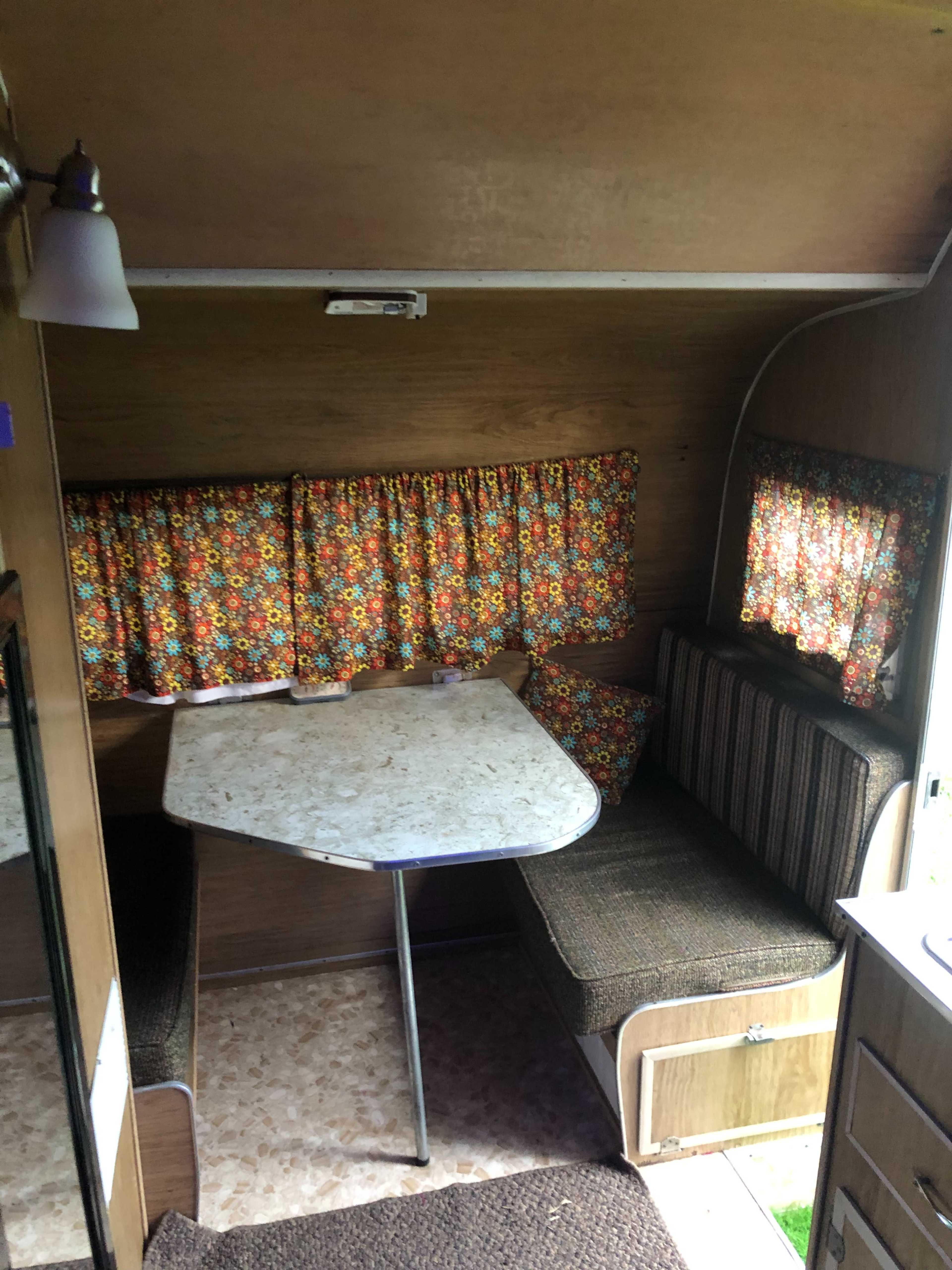The interior of a vintage camper features a table with a triangular shape, surrounded by cushioned benches and floral-patterned curtains.