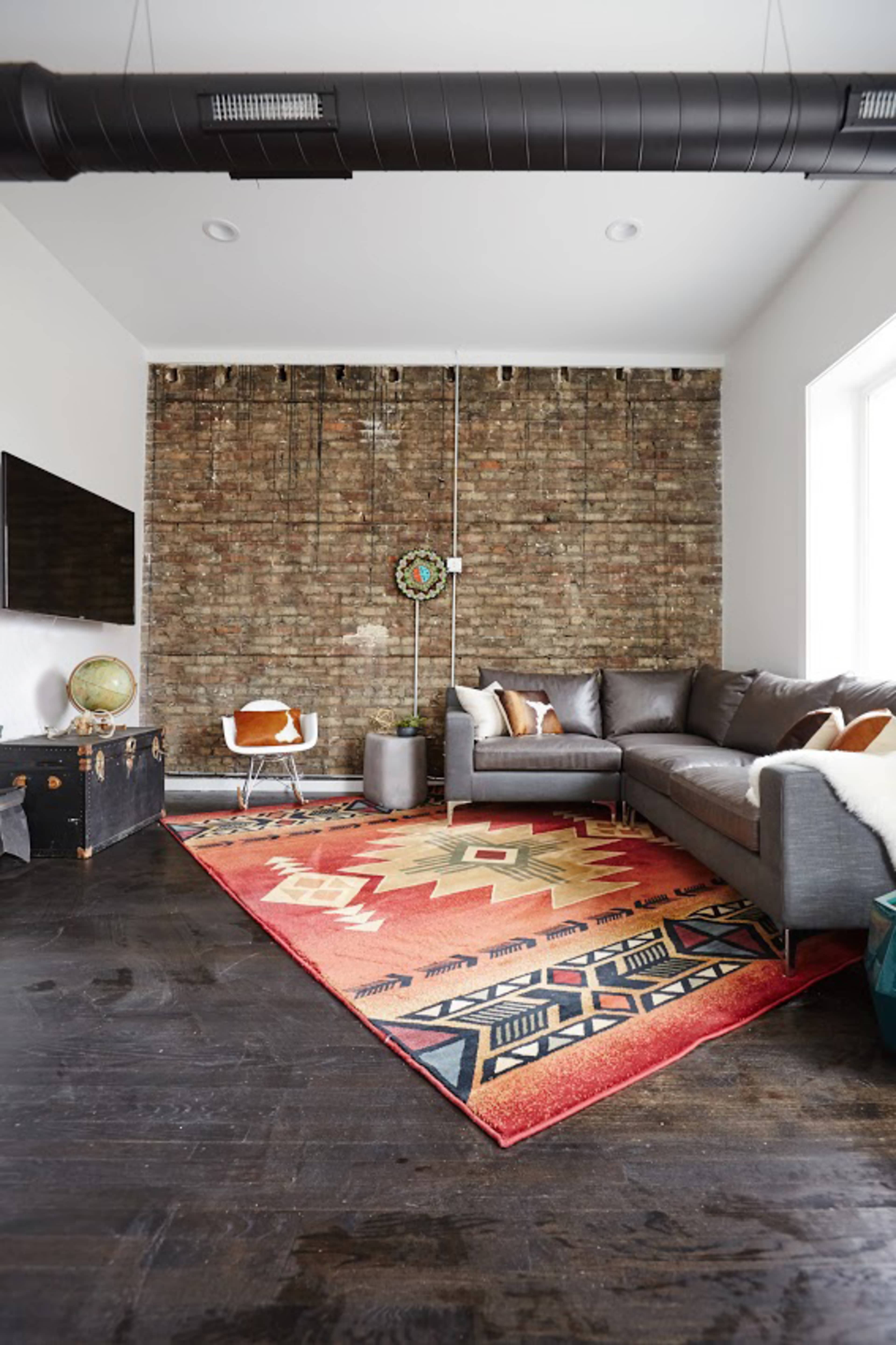 A living room with a sectional gray sofa, a patterned area rug, a brick accent wall, and a flat-screen TV mounted on the wall.