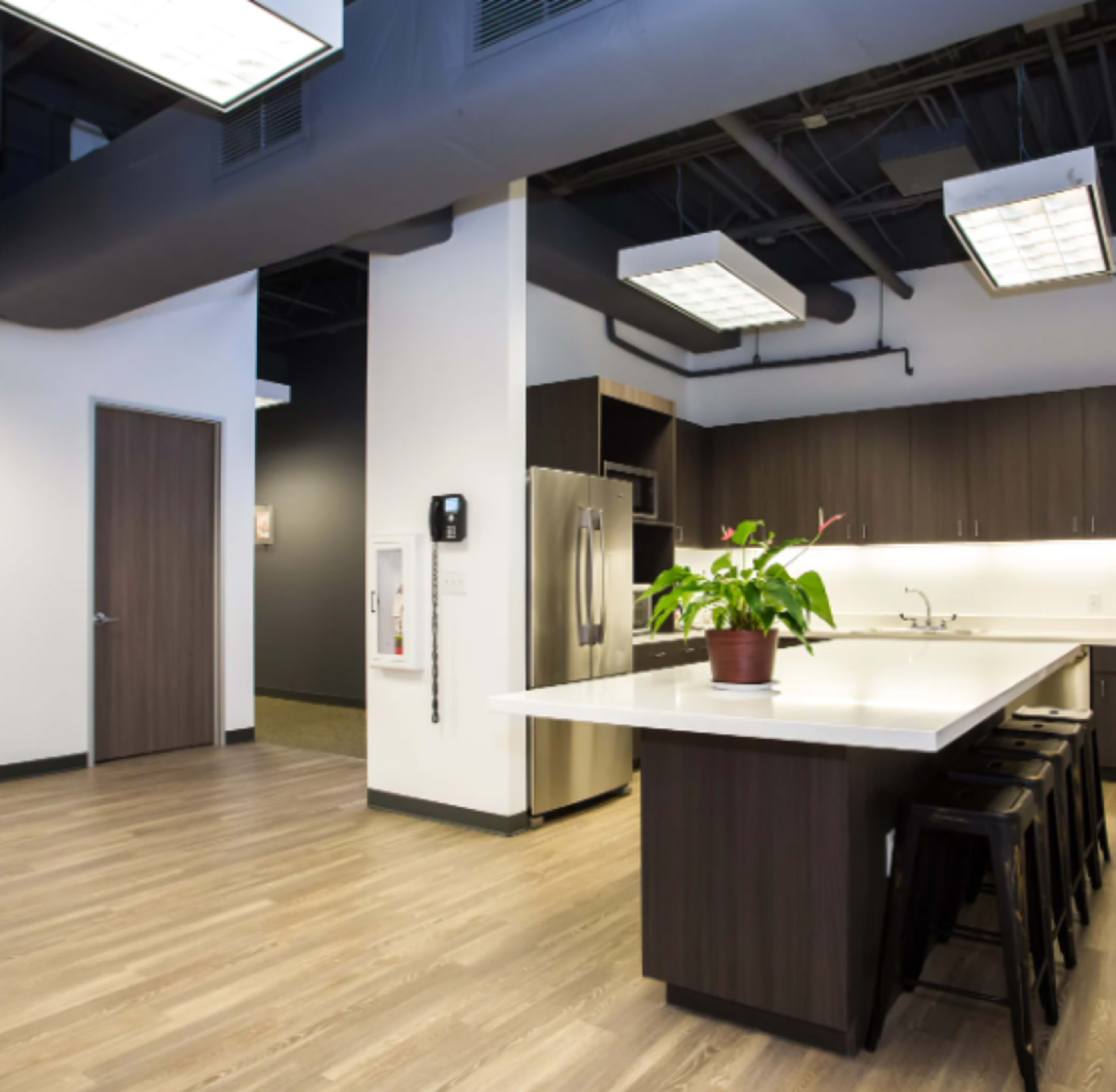 The image shows a modern kitchen area within an office, featuring a large island with seating, stainless steel appliances, and dark cabinetry.