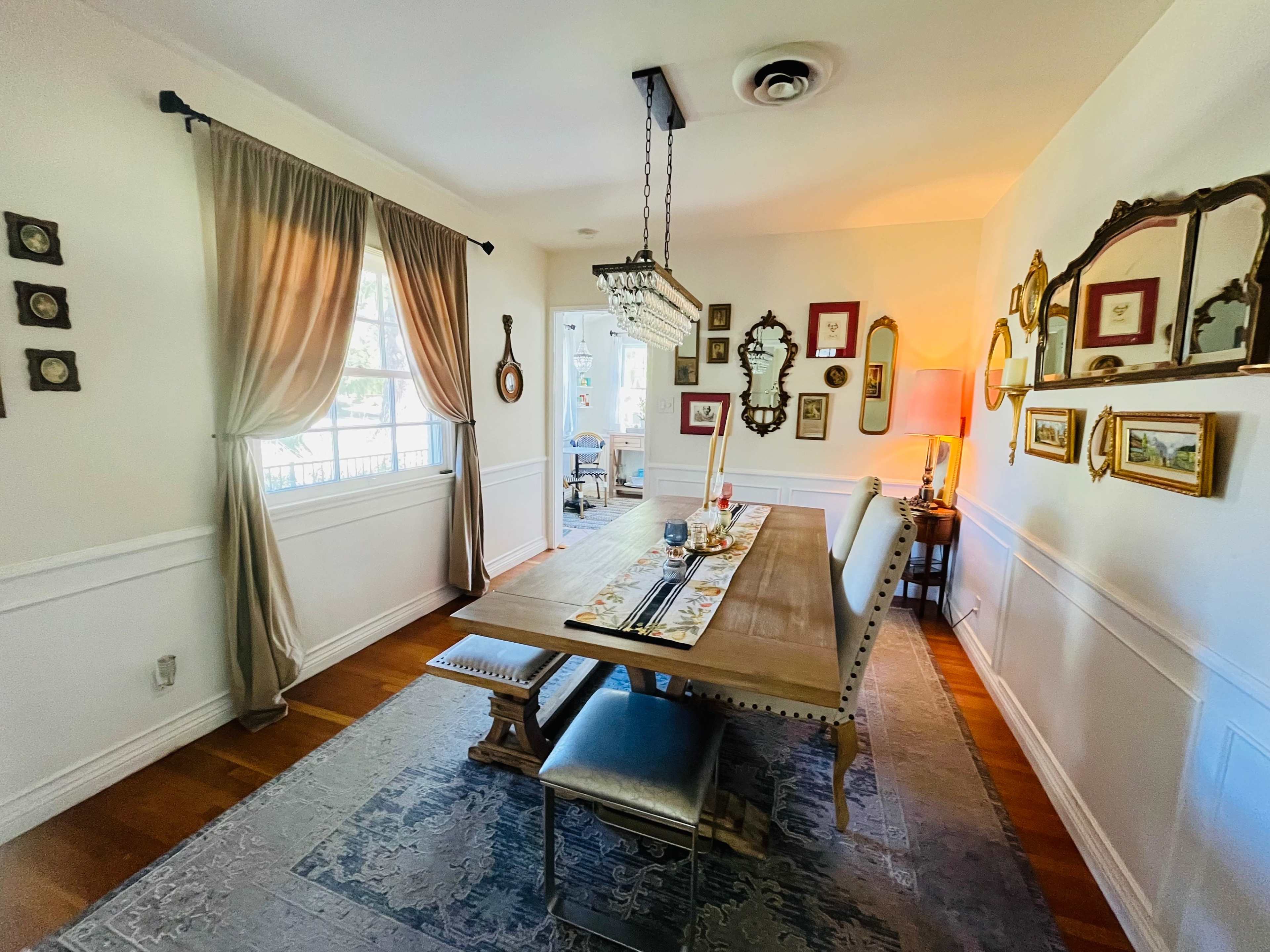 A spacious dining room features a long wooden table surrounded by upholstered chairs, with decorative mirrors and framed artwork on the walls.