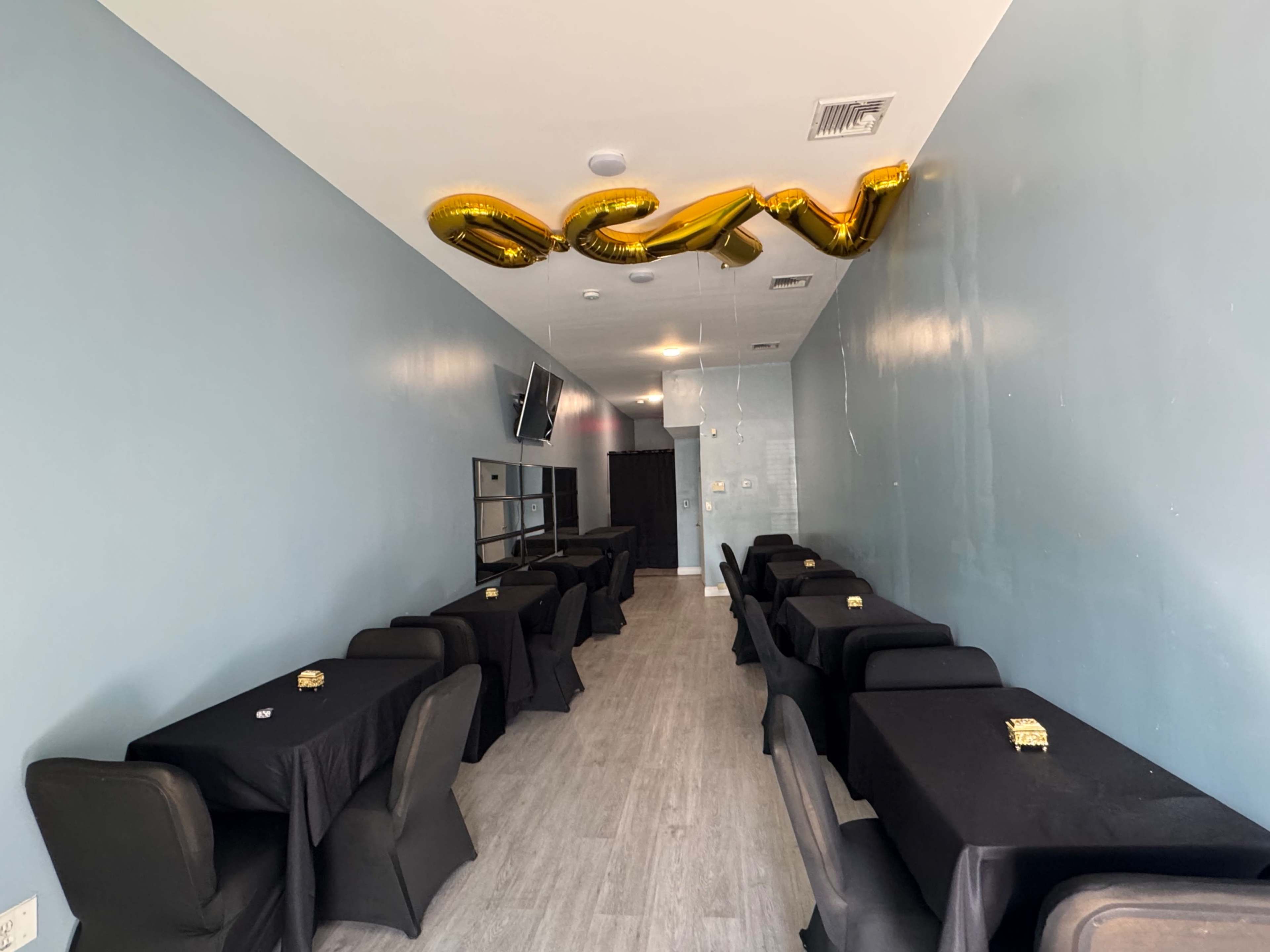 The image shows a narrow dining area with several black tables covered with tablecloths and gold balloons spelling "OPEN" hanging from the ceiling.