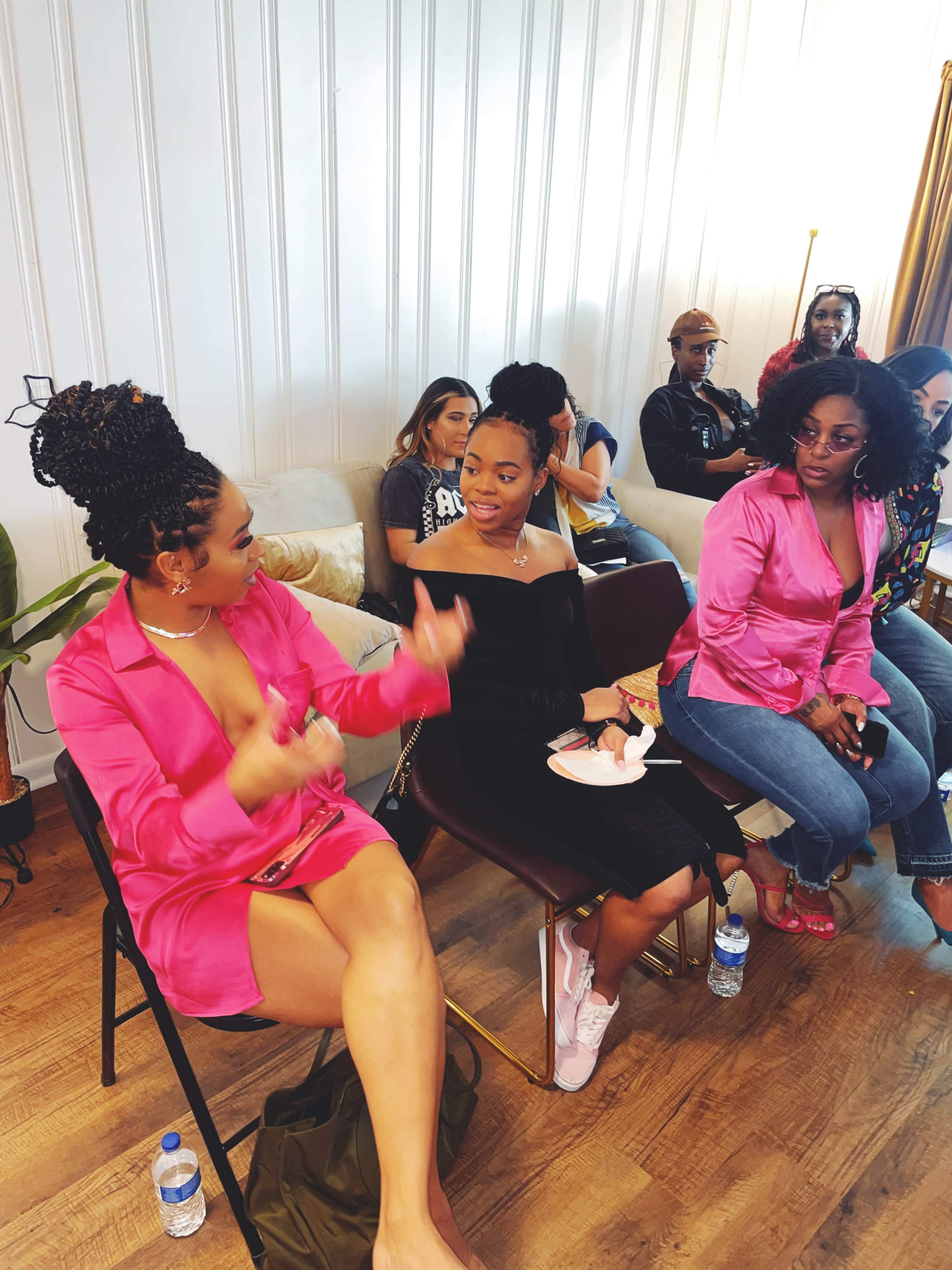 A group of people sits in a modern, well-lit room, with two women engaged in conversation while others observe.
