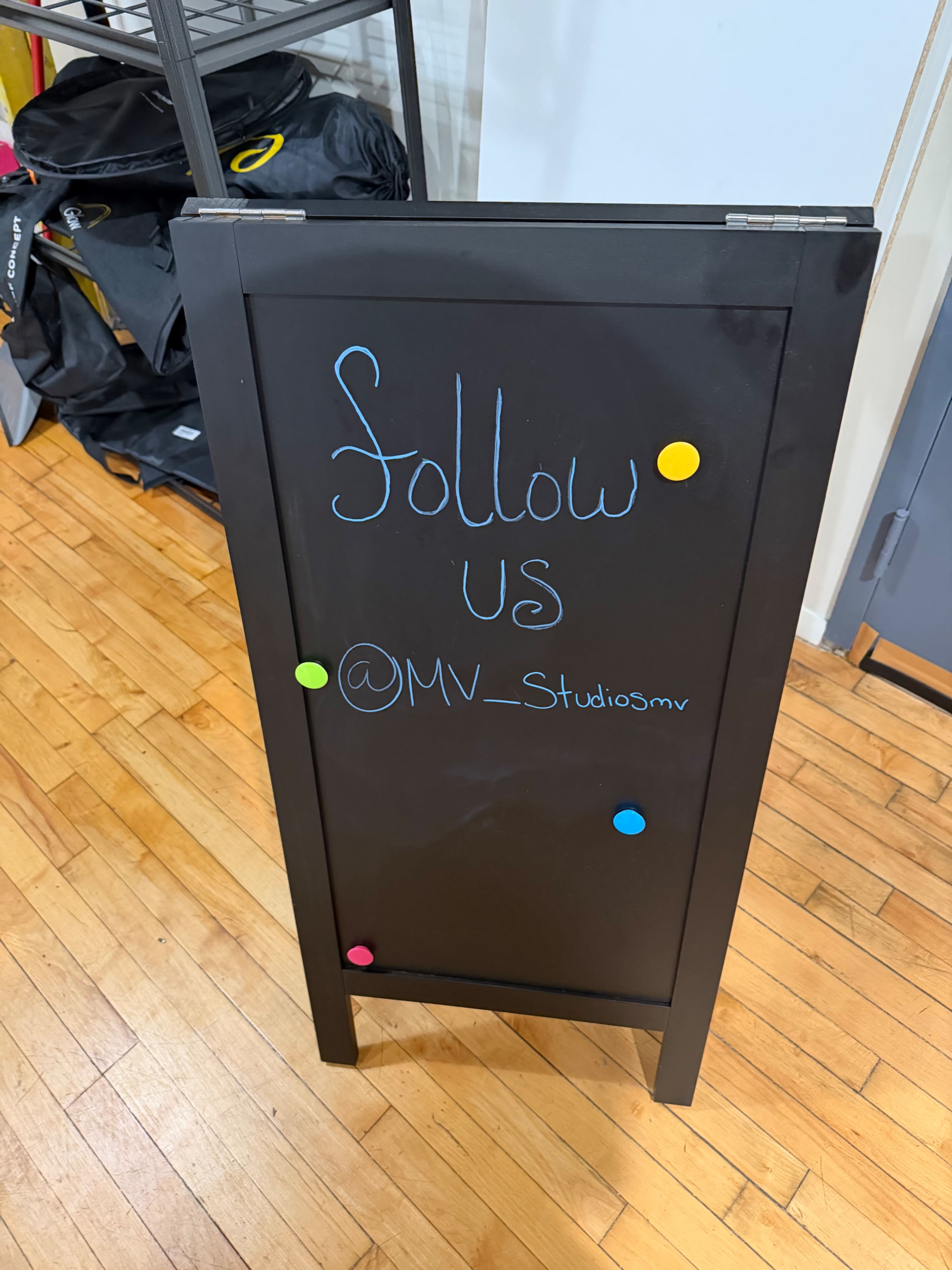 A black chalkboard sign displays the message "Follow Us @MV_Studiosmv" and is attached to a wooden floor in a studio setting.