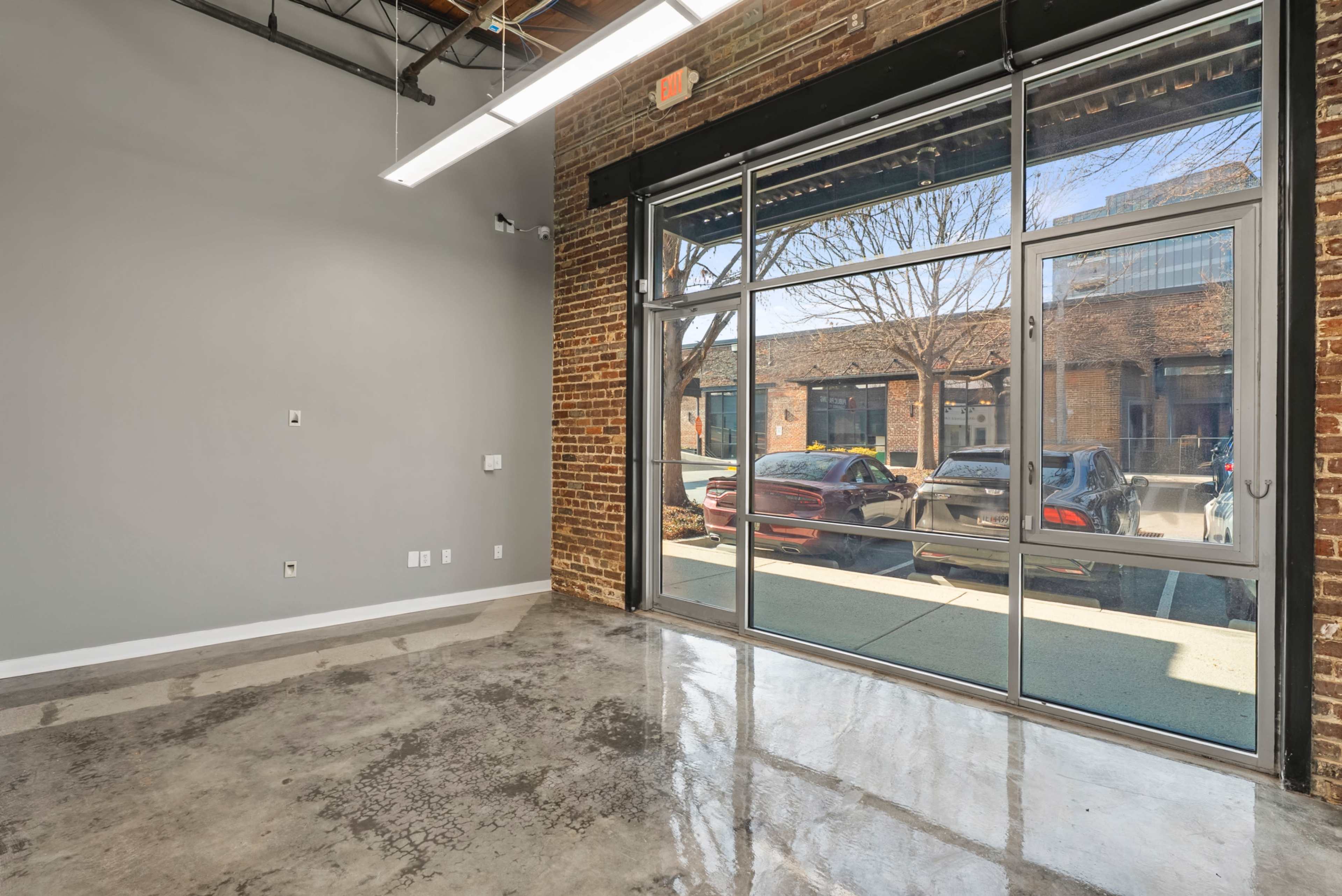 The image shows an empty industrial space with exposed brick walls, large glass doors, and polished concrete flooring.