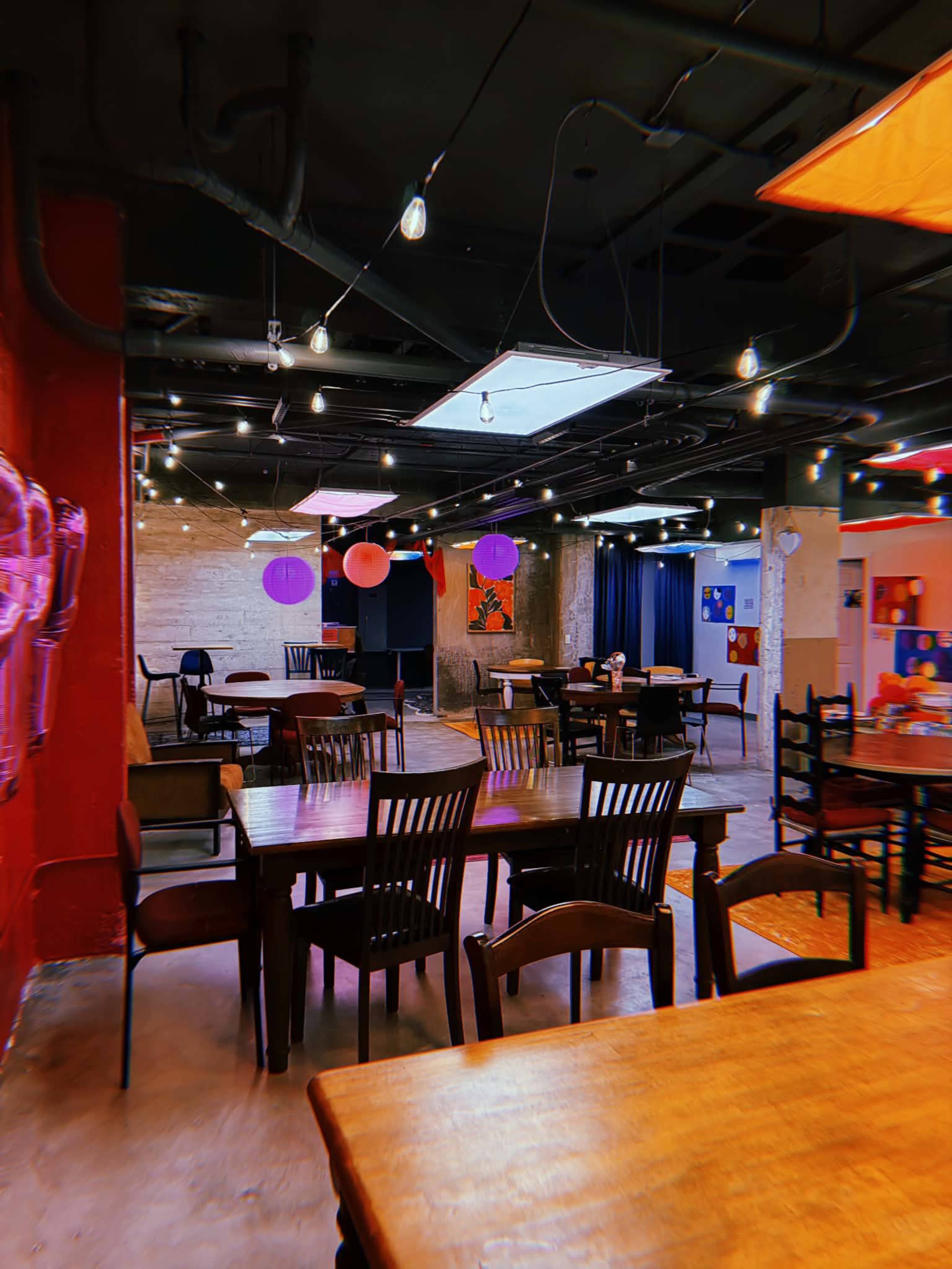 The image shows a large, modern restaurant interior with wooden tables, chairs, and colorful decorations hanging from the ceiling.