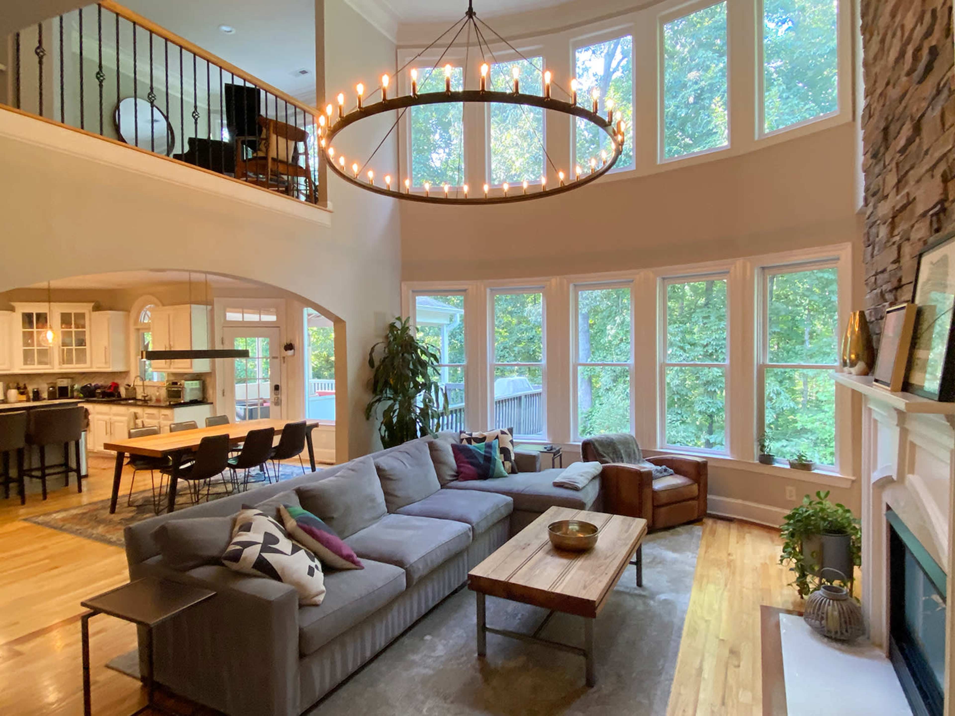 A spacious living room with large windows, a circular chandelier, a gray sectional sofa, a wooden coffee table, and a stone fireplace, overlooking a wooded area.