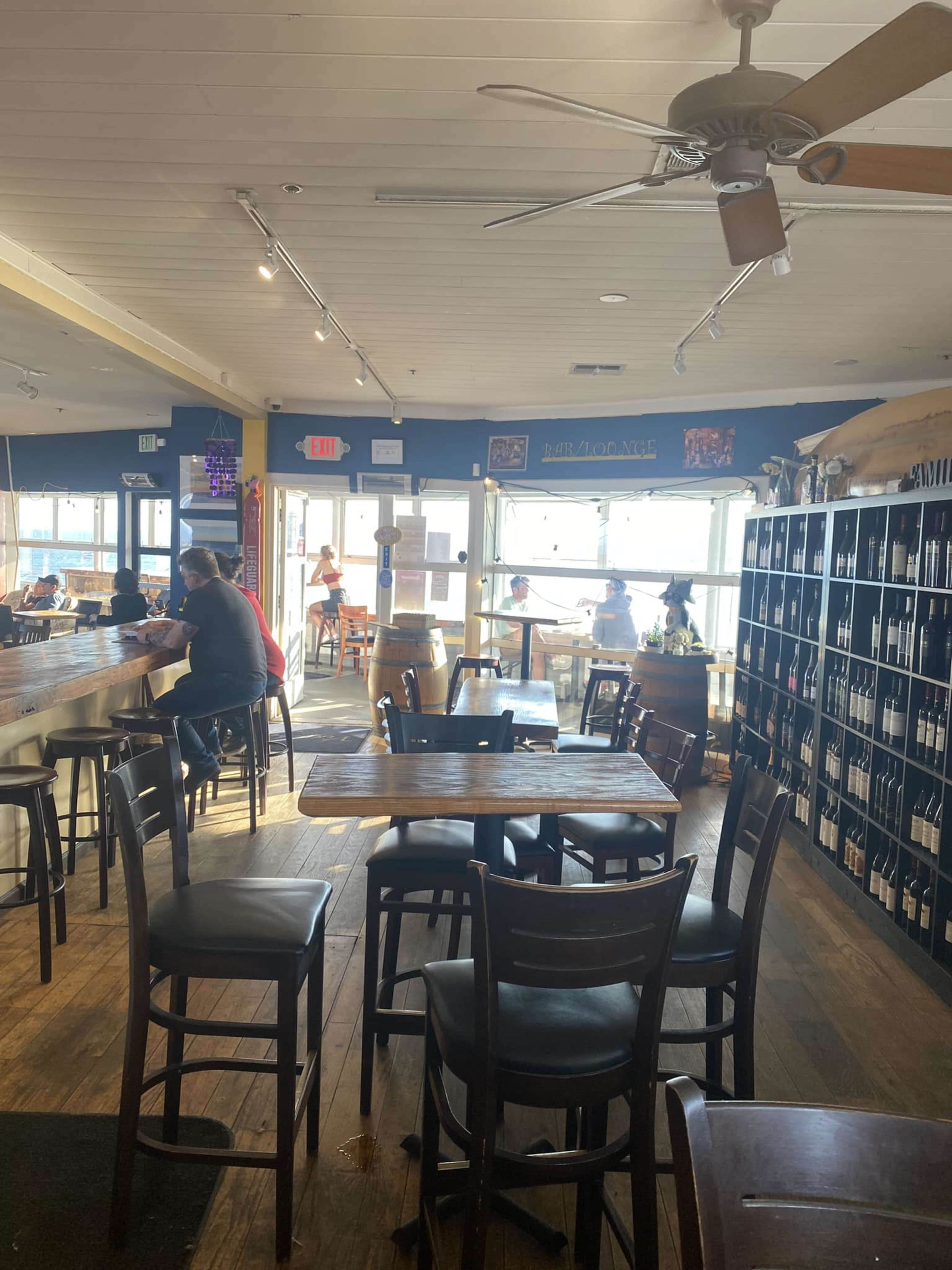 The image shows the interior of a restaurant with wooden tables and chairs, a bar area, and large windows facing an outdoor seating area.