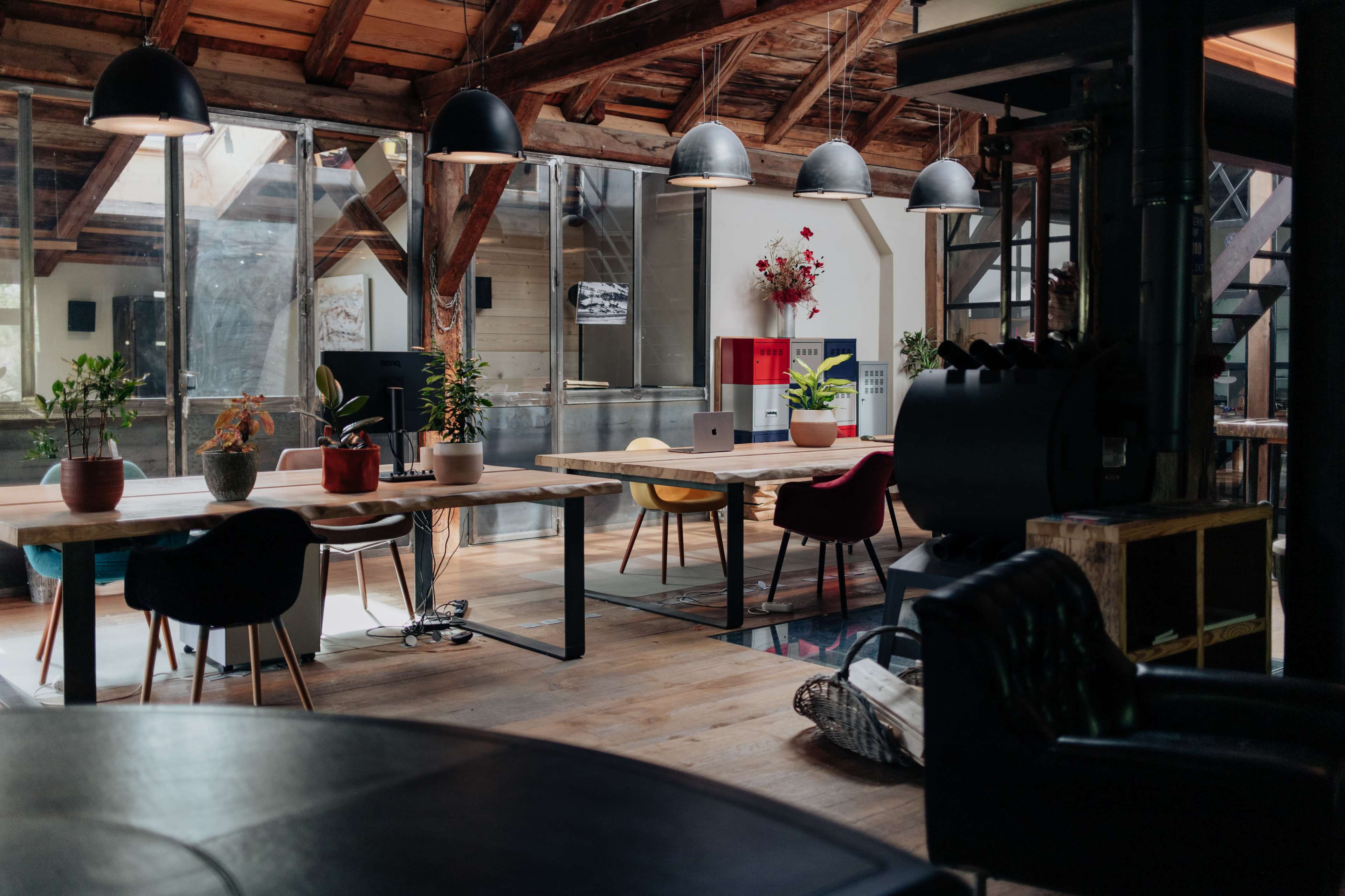 The image shows a spacious, modern workspace with wooden tables, various chairs, plants, and large windows letting in natural light.