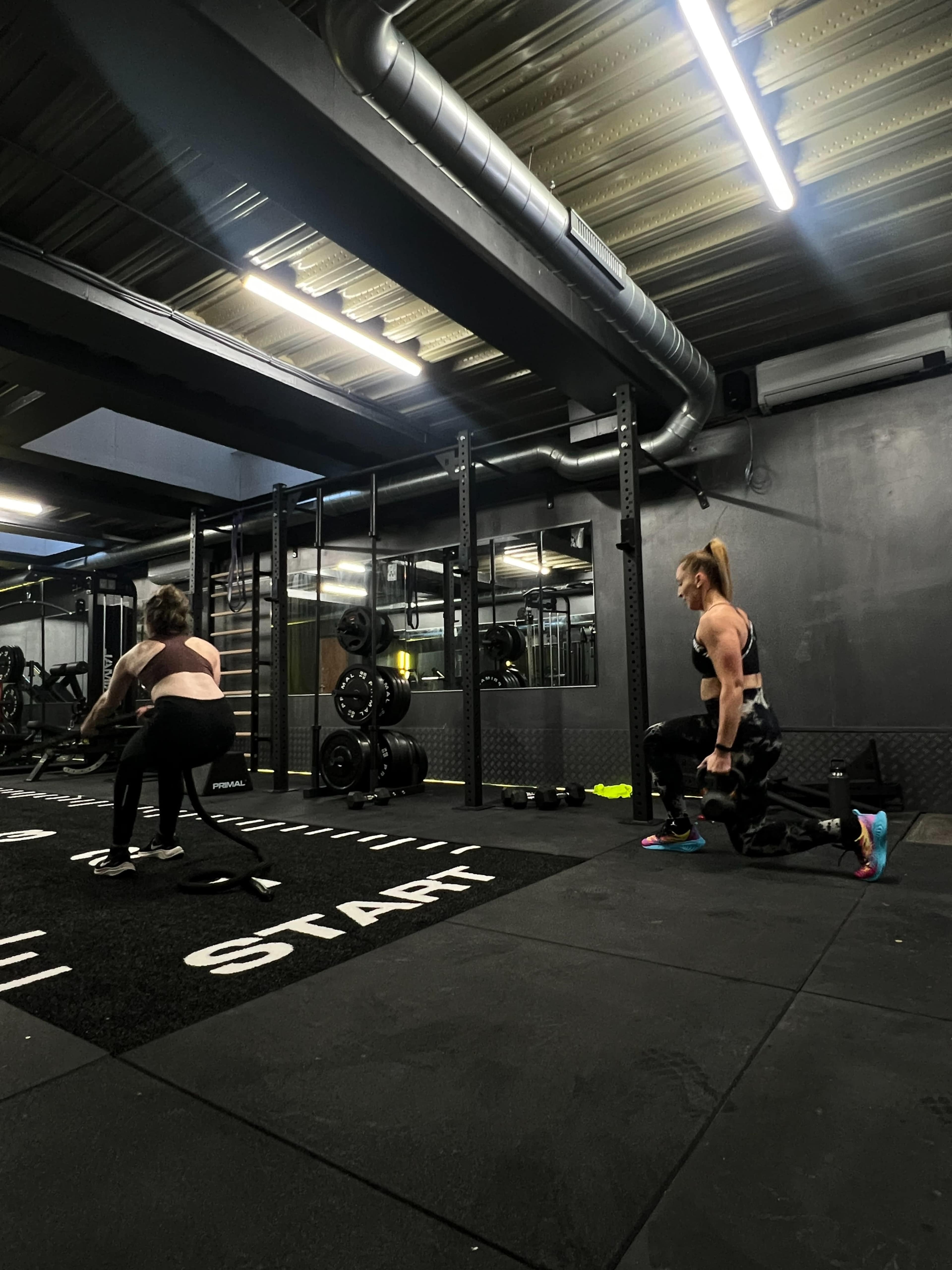 Two individuals are exercising in a gym, one using battle ropes and the other performing a lunge, with workout equipment visible in the background.