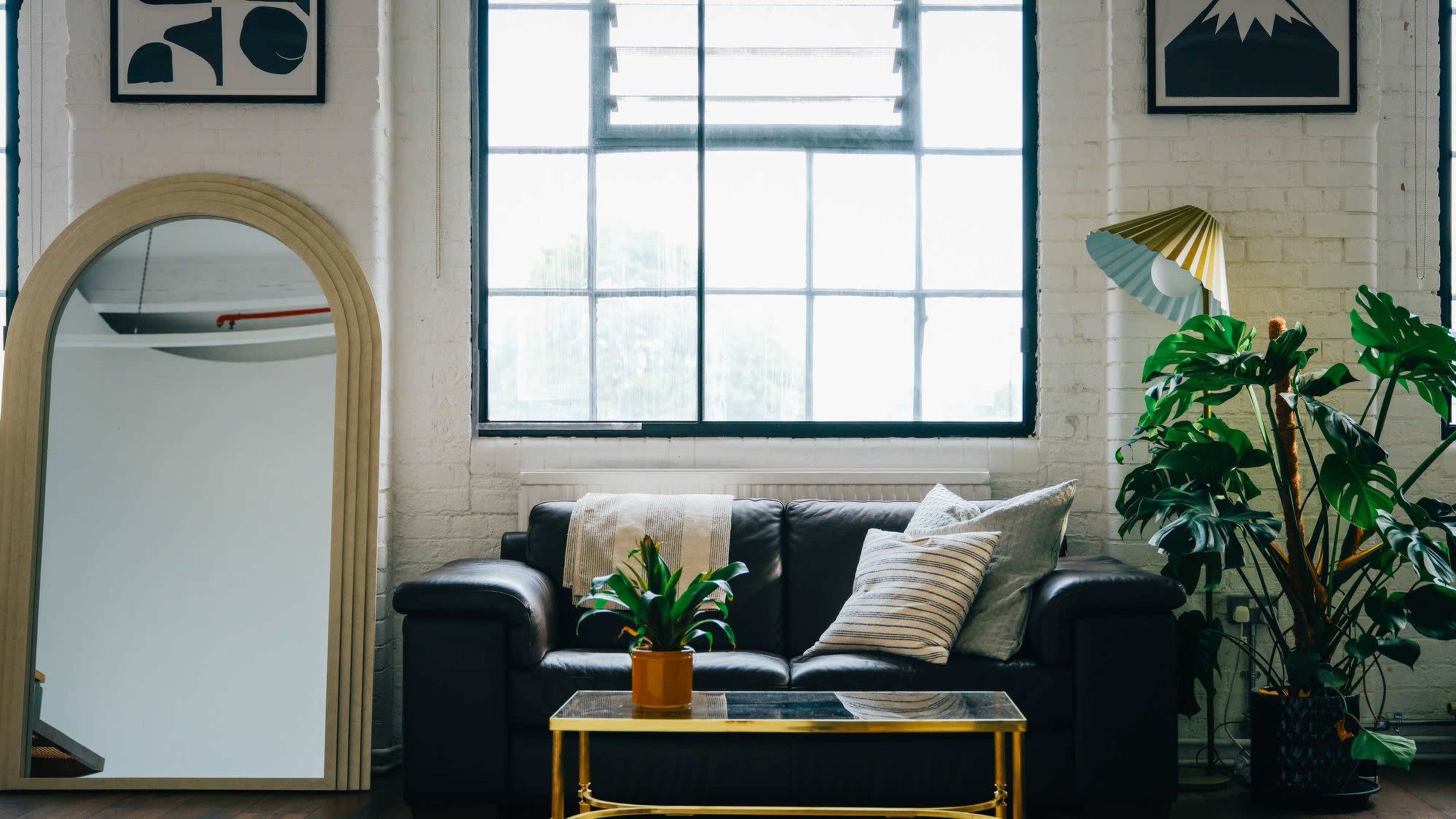 The image shows a living room featuring a black leather sofa, a gold coffee table, a large mirror, a floor lamp, and several indoor plants, all arranged near a large window.