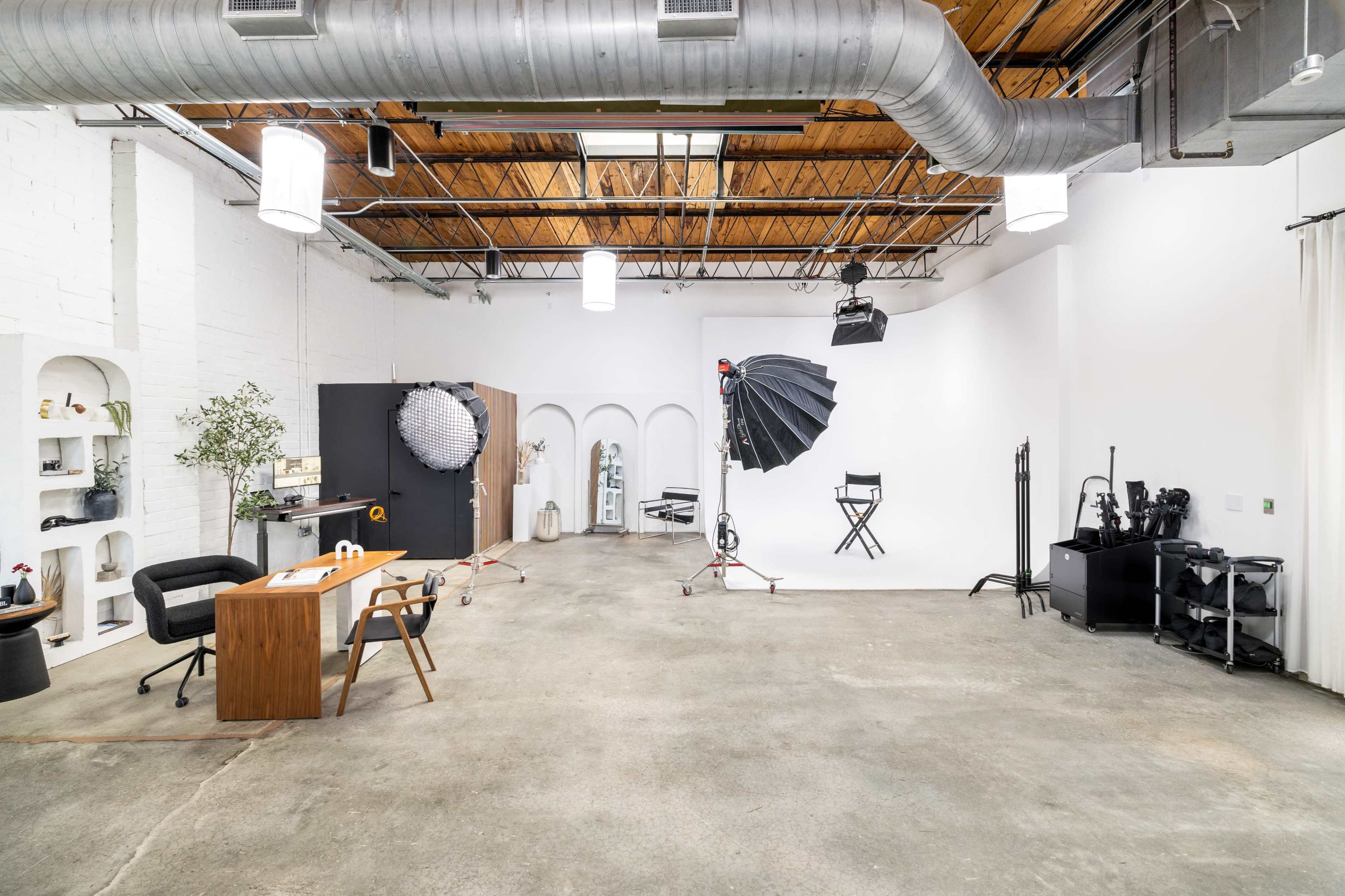 A spacious photography studio featuring various equipment, a desk with a chair, and a backdrop setup with lighting.