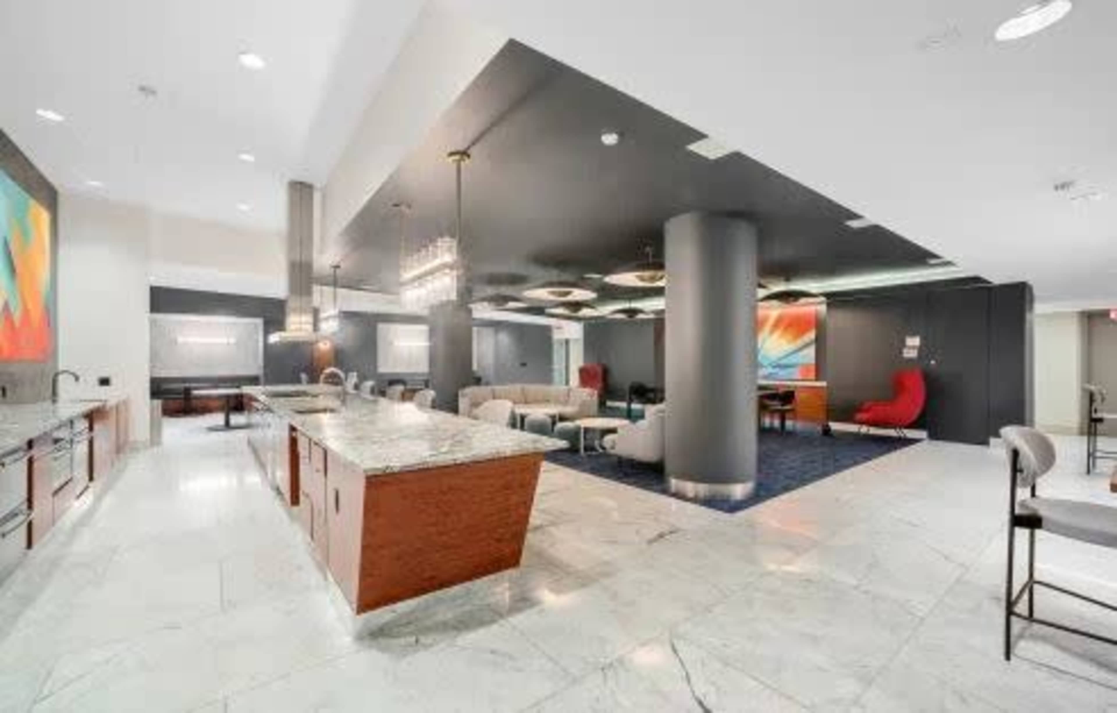 The image shows a modern lobby area with a large kitchen island, seating arrangements, and abstract artwork on the walls.