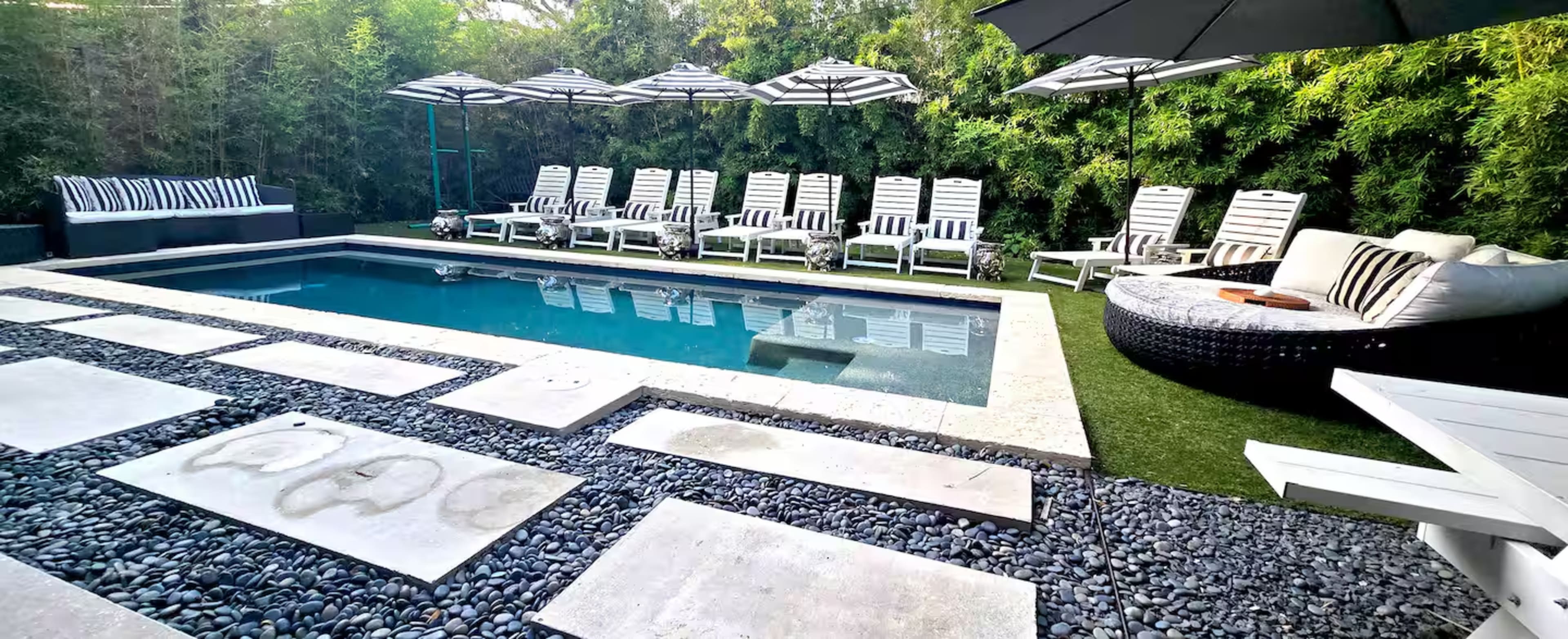 The image shows a backyard area featuring a swimming pool surrounded by striped lounge chairs, umbrellas, and stone pathways lined with pebbles.