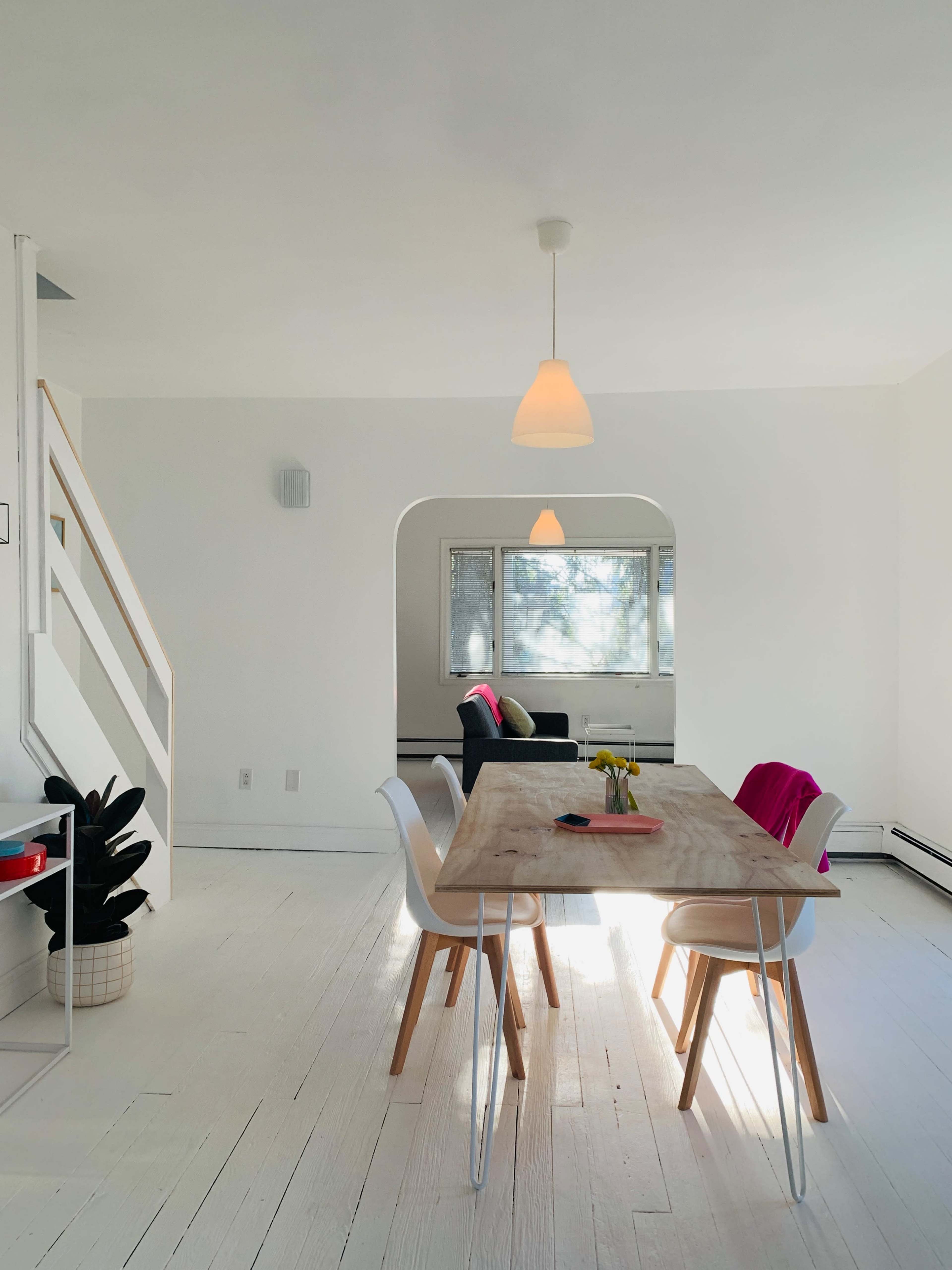 A bright dining area features a wooden table surrounded by white chairs, with a staircase visible in the background and a couch in a cozy corner.