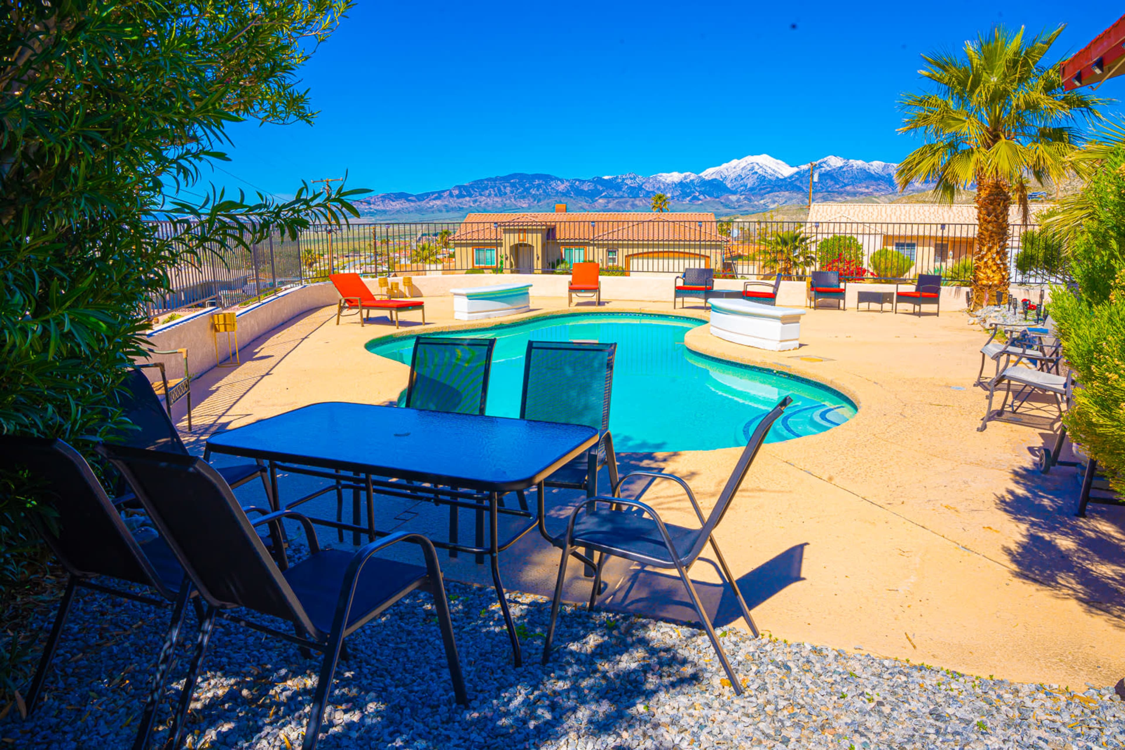 A pool area surrounded by lounge chairs and a table, with mountains in the background and clear blue skies.