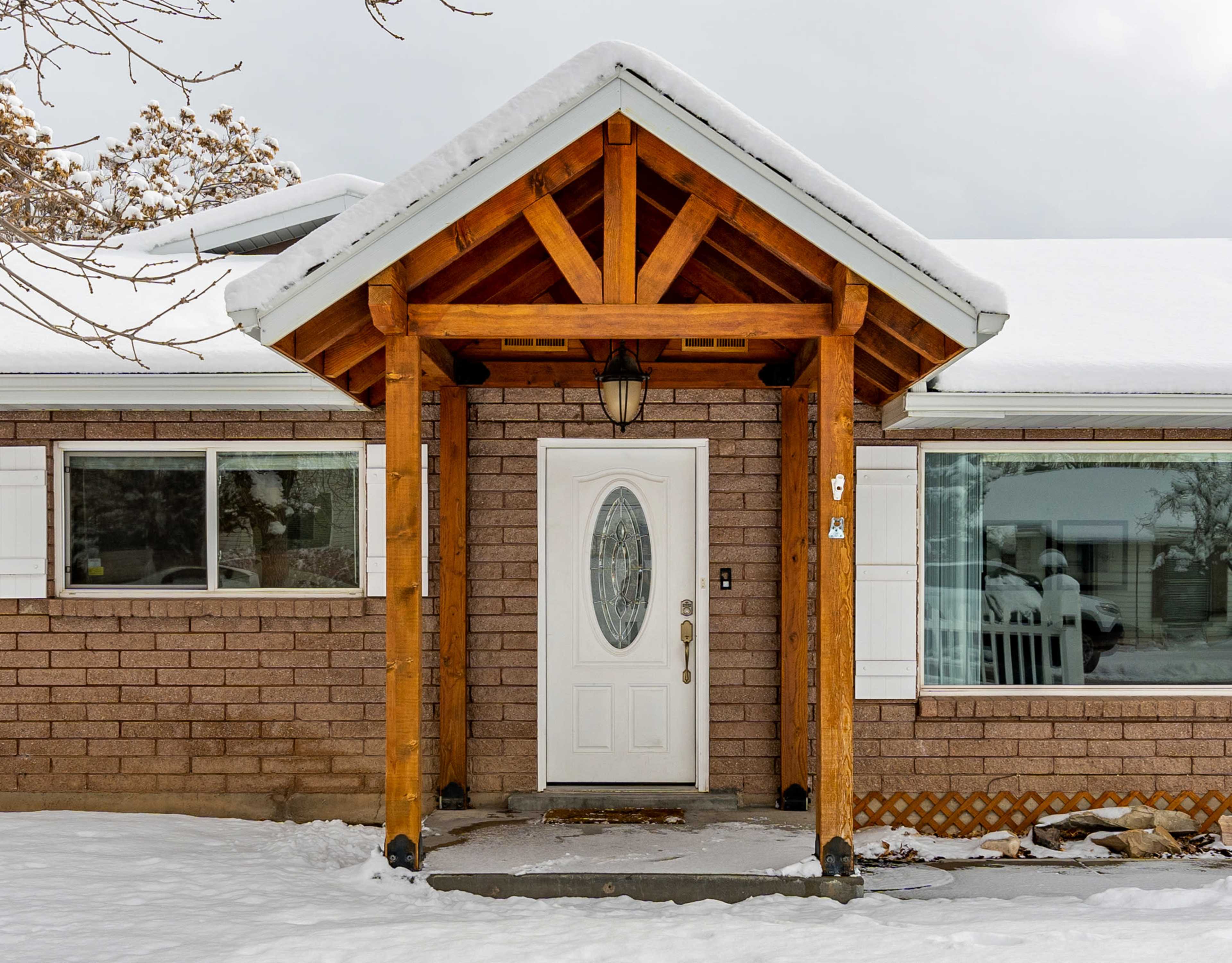 A front entrance of a house, featuring a wooden canopy and a white door, surrounded by a layer of snow.