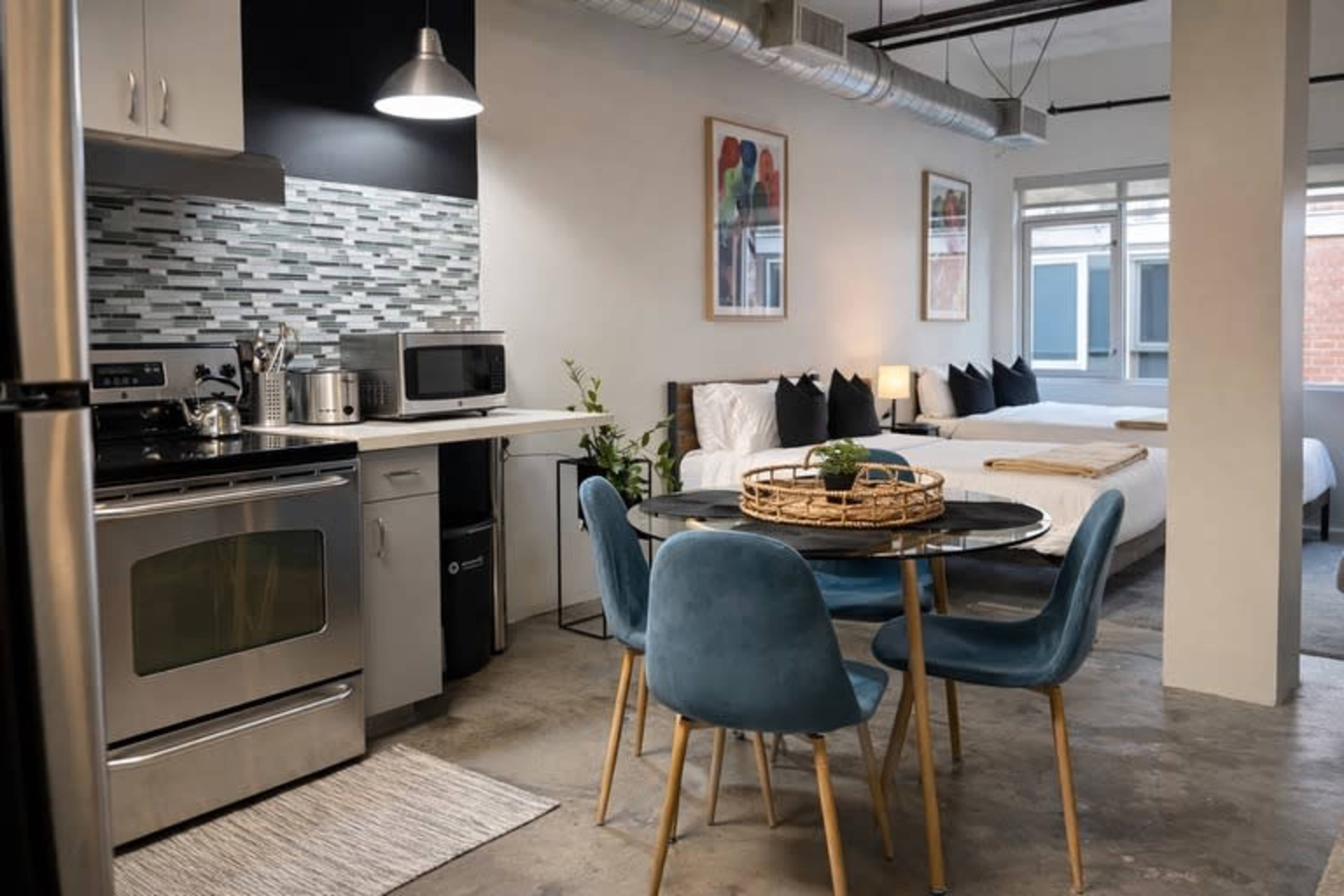 The image shows a modern studio apartment featuring a kitchen area with stainless steel appliances and a dining table with blue chairs, alongside a sleeping area with two beds.