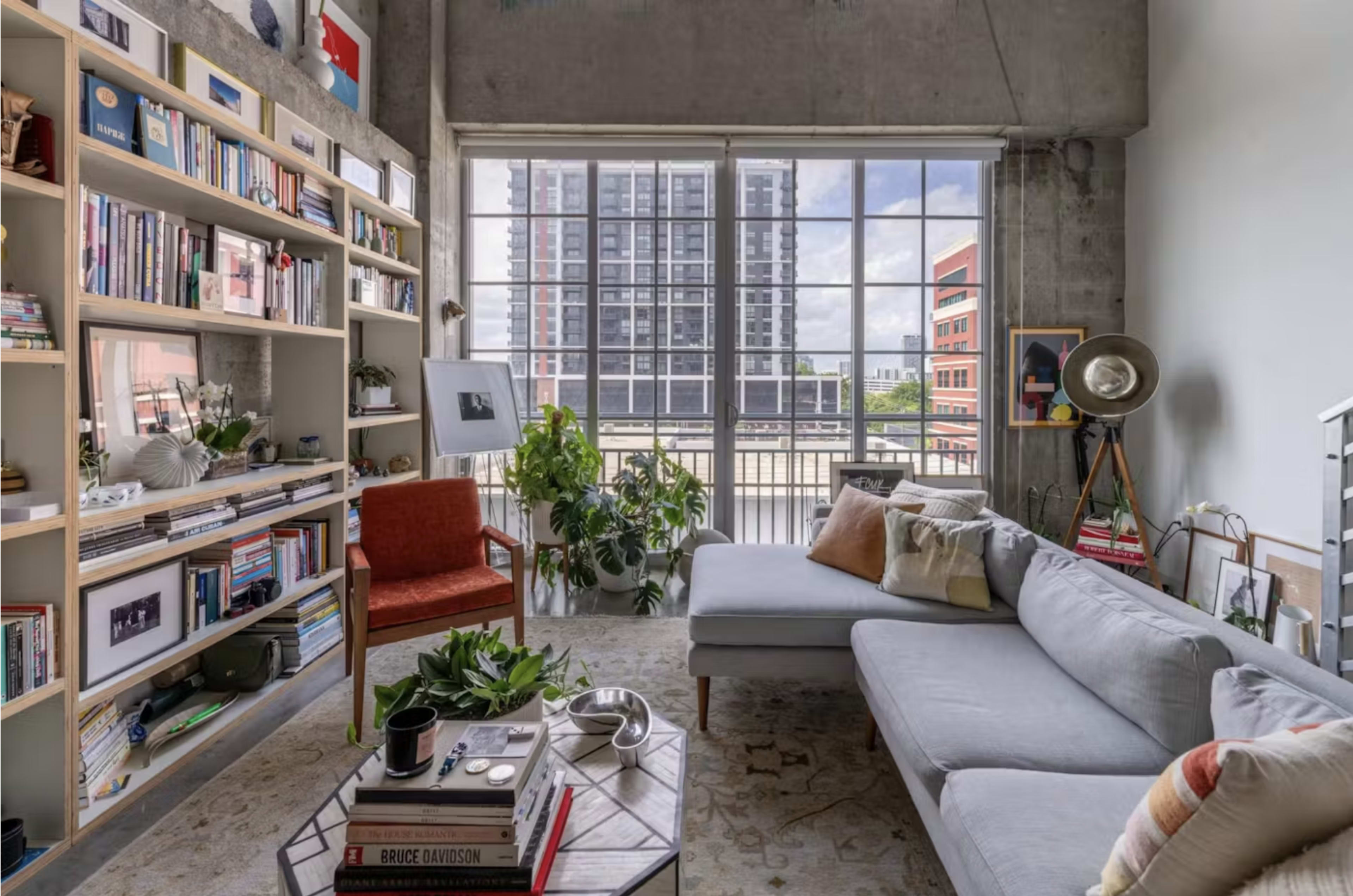 A modern living room features a light gray sectional sofa, shelves filled with books and decor, plants, and large windows showcasing a city view.