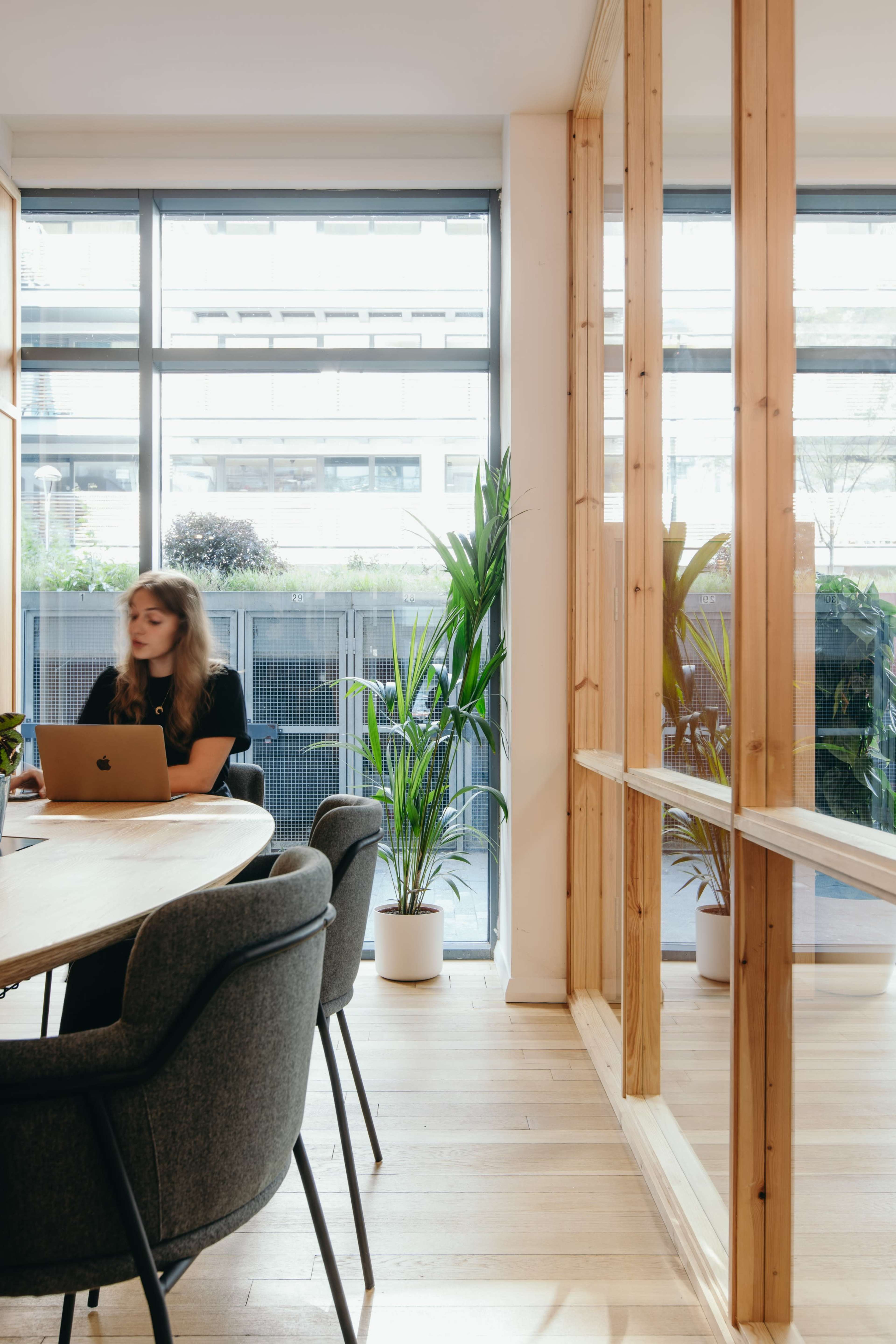 A woman works on a laptop at a wooden table in a modern office with large windows and potted plants.