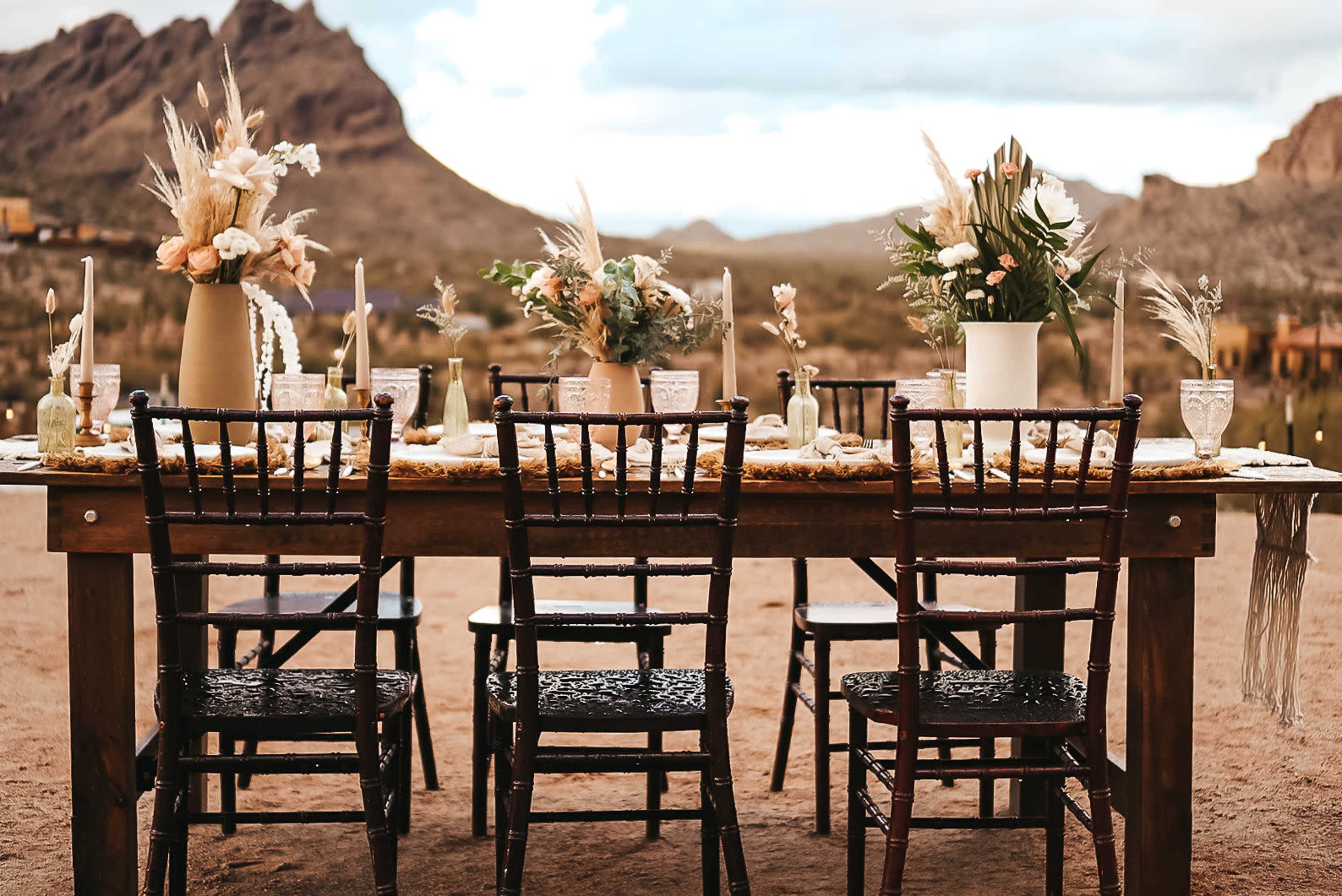 A rustic outdoor dining table is set with floral arrangements, candles, and chairs, surrounded by desert mountains.