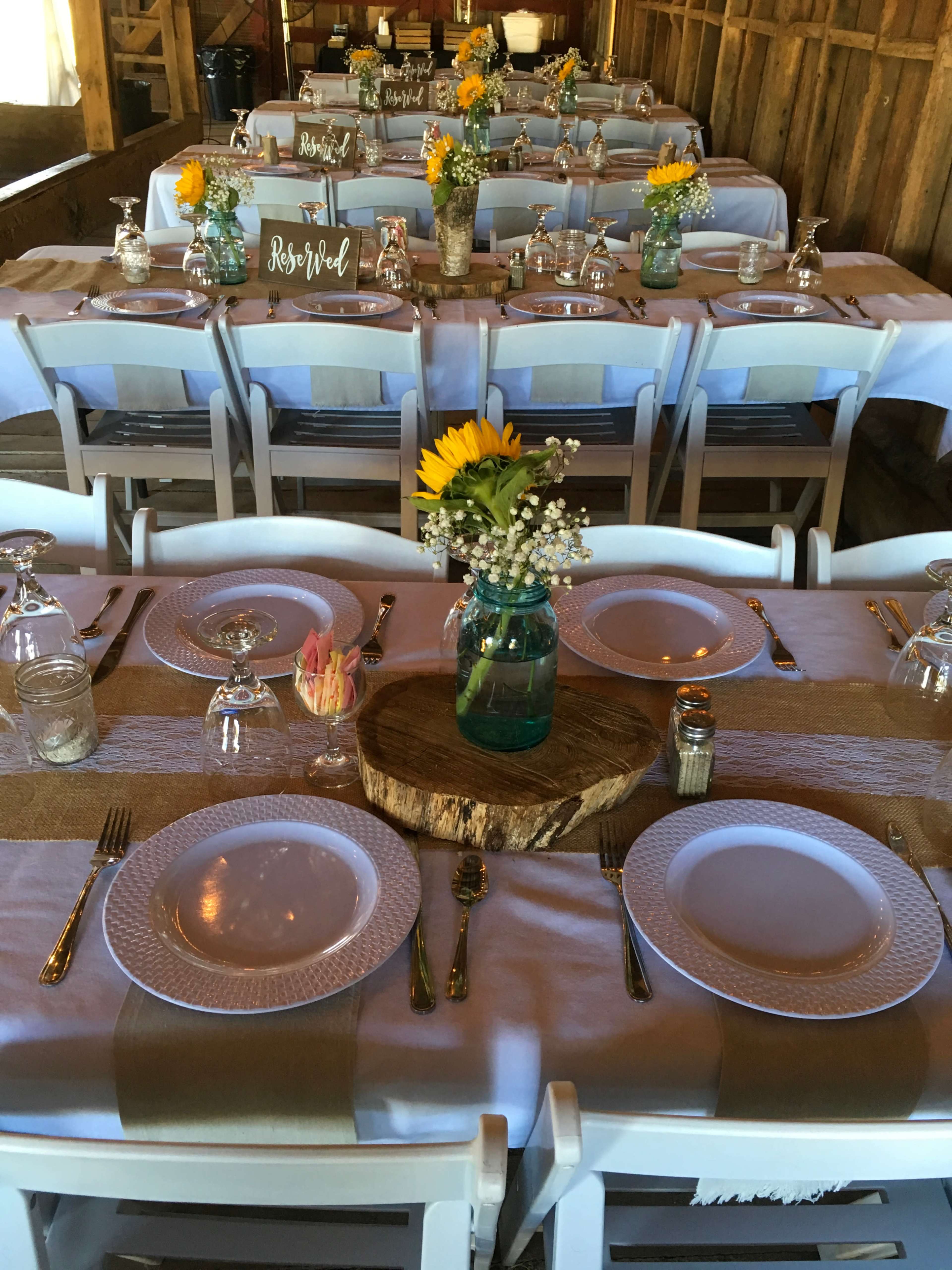 A wooden table is set for a meal in a rustic barn, featuring white plates, mason jars with flowers, and neatly arranged utensils.
