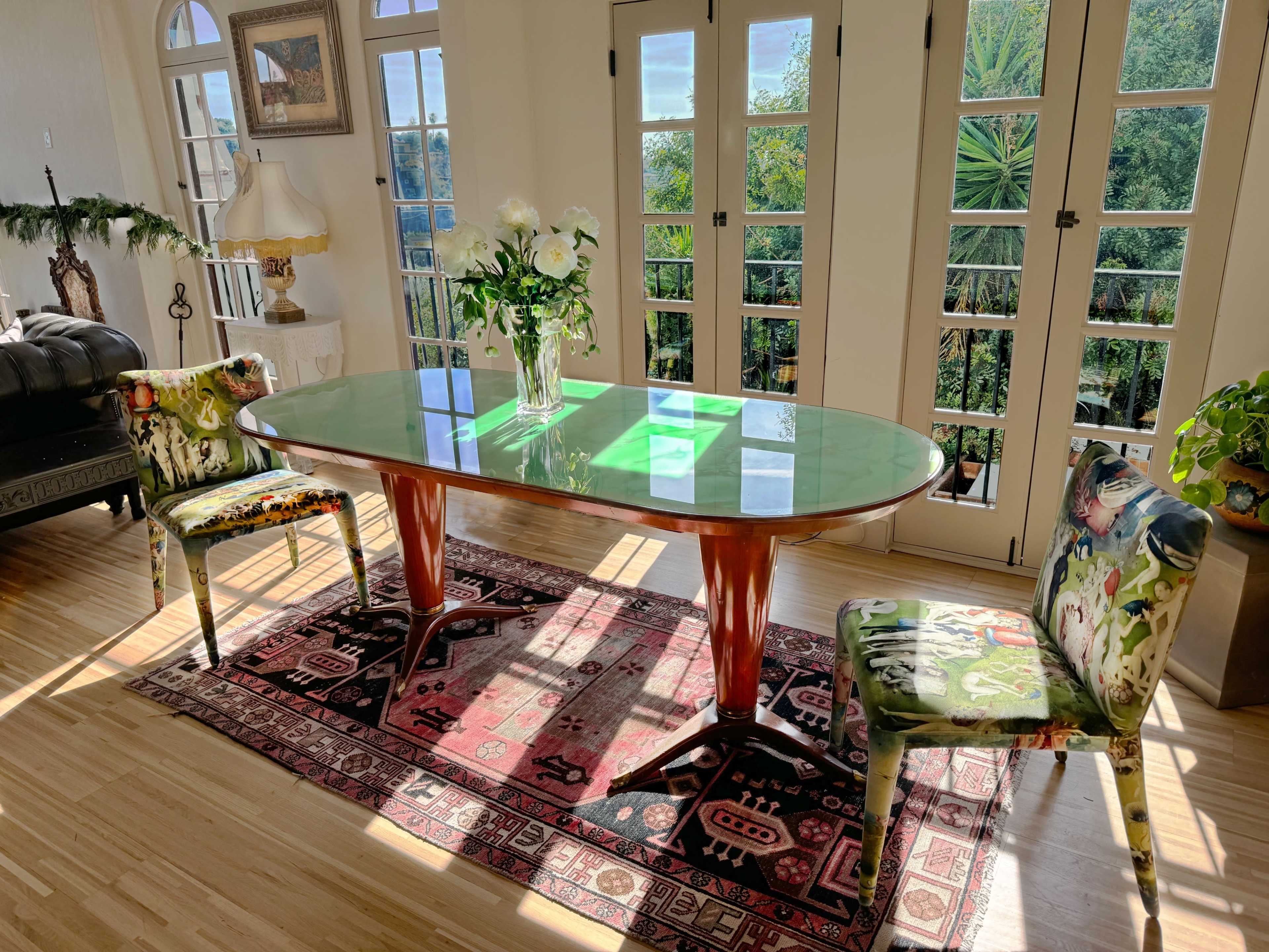 A dining table with a green glass top is surrounded by two patterned chairs on a colorful rug, with large windows allowing sunlight to enter the room.