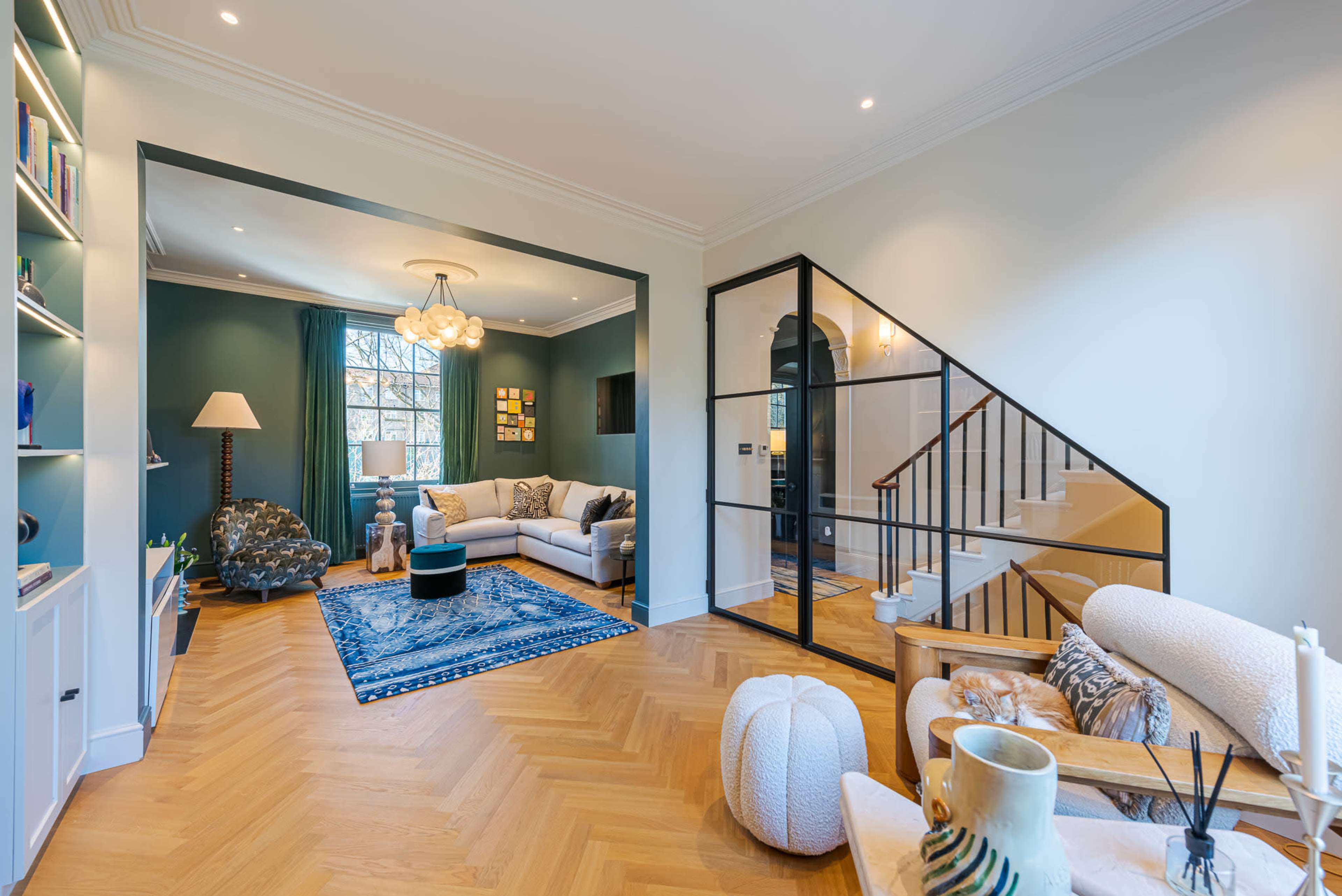 The image shows a modern living room with a blue patterned rug, a large window, and a staircase leading to an upper level.