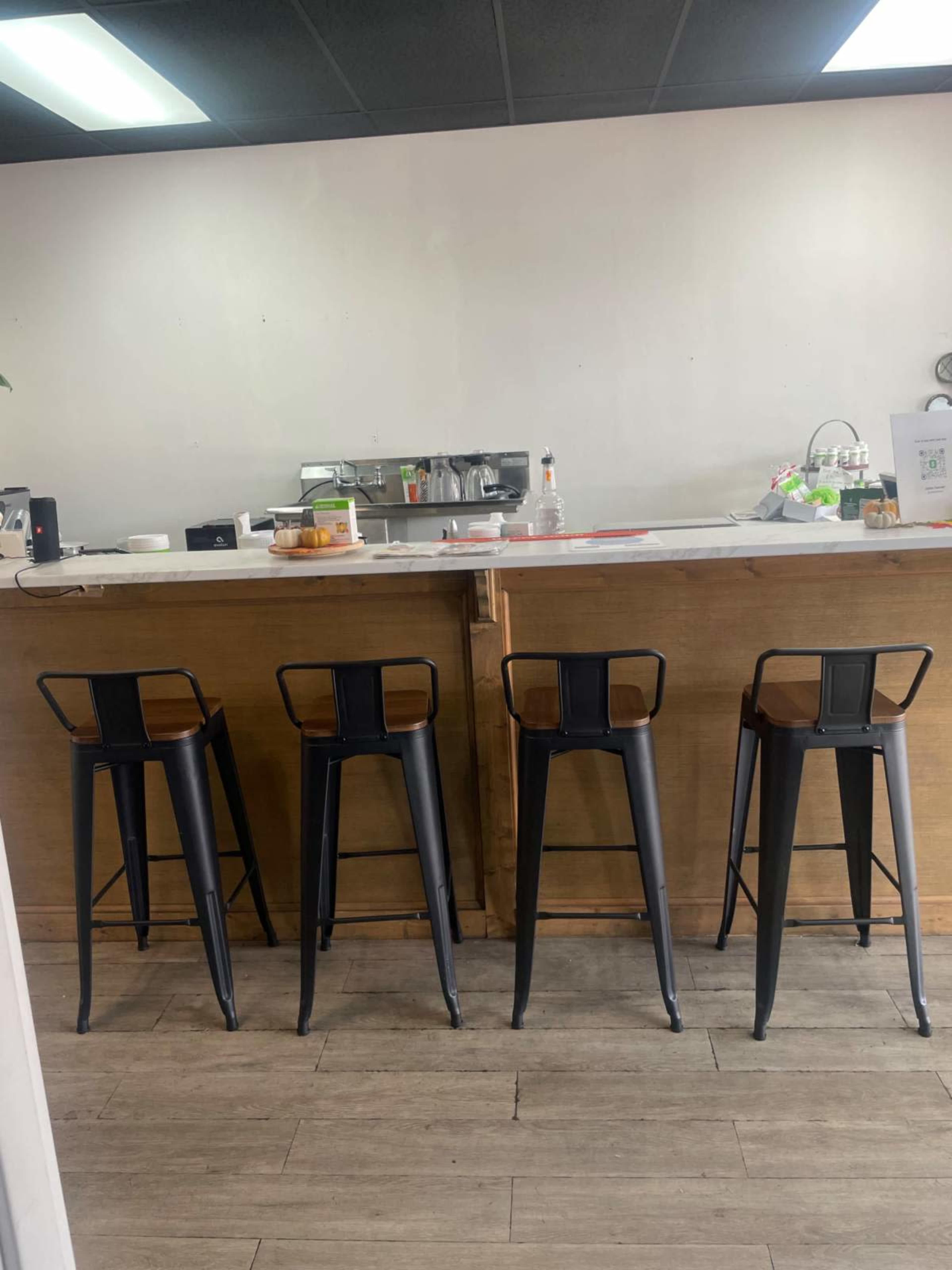 The image shows a minimalistic cafe counter with four black stools lined up in front of a wooden bar.