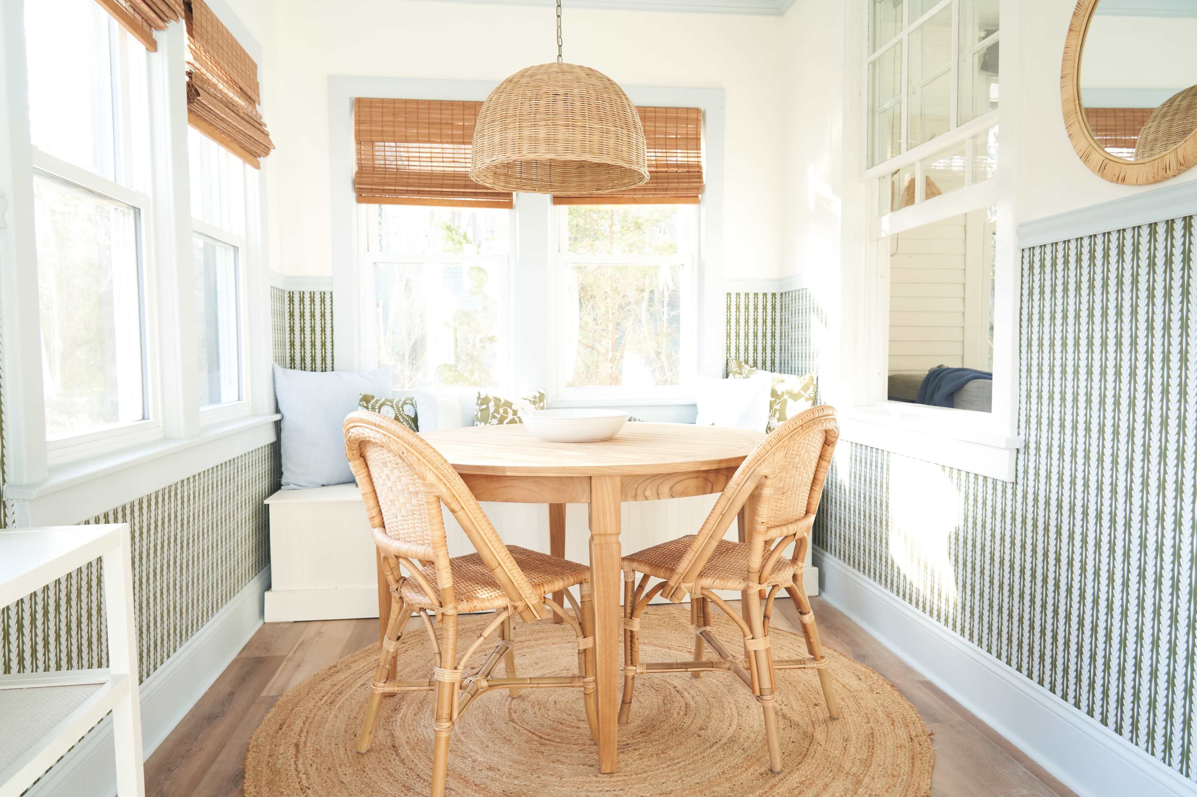 A round wooden table with four wicker chairs is centered in a sunlit dining area featuring large windows and patterned wallpaper.