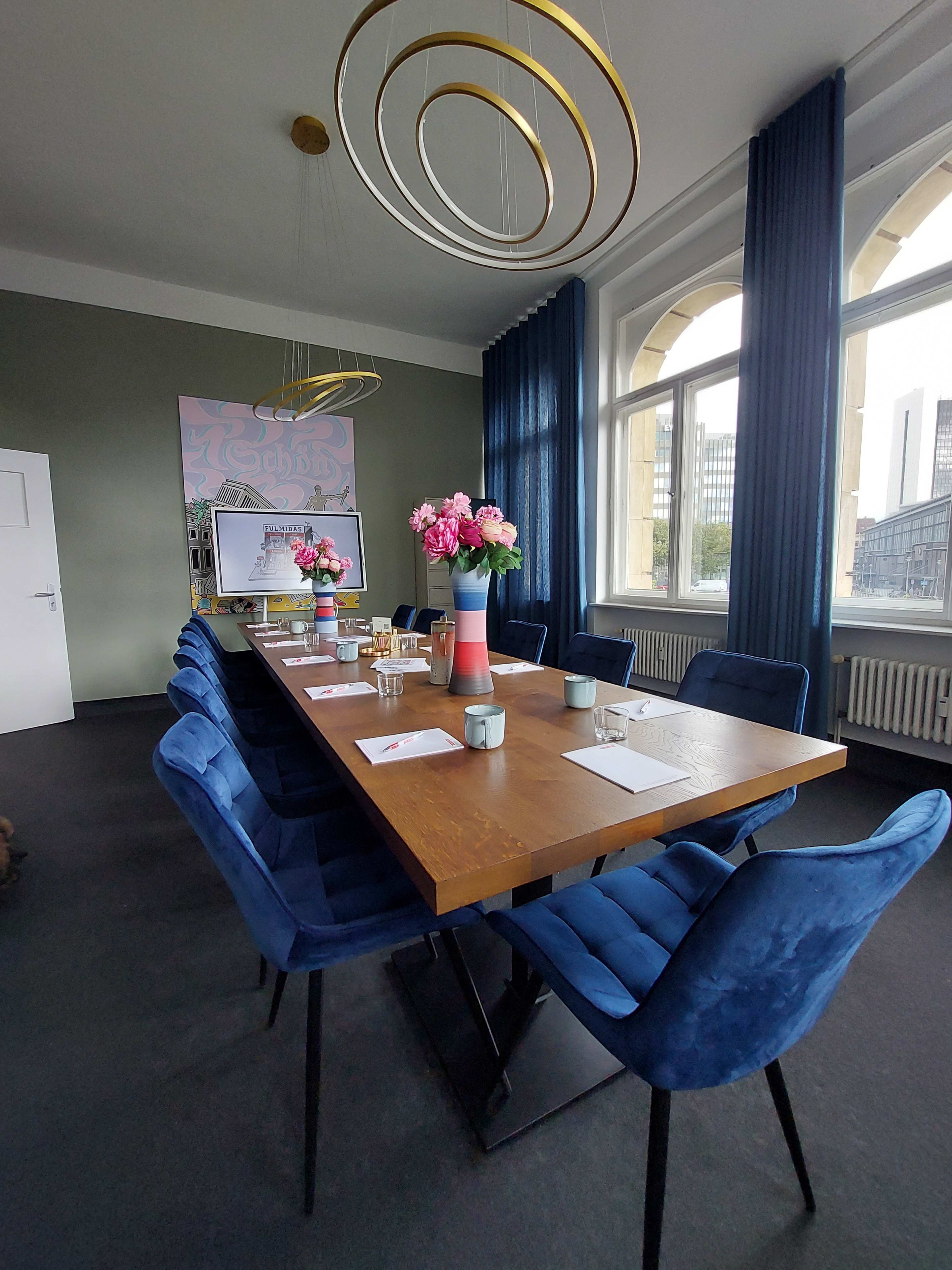 A conference room features a long wooden table surrounded by blue chairs, with floral centerpieces and large windows draped with curtains.