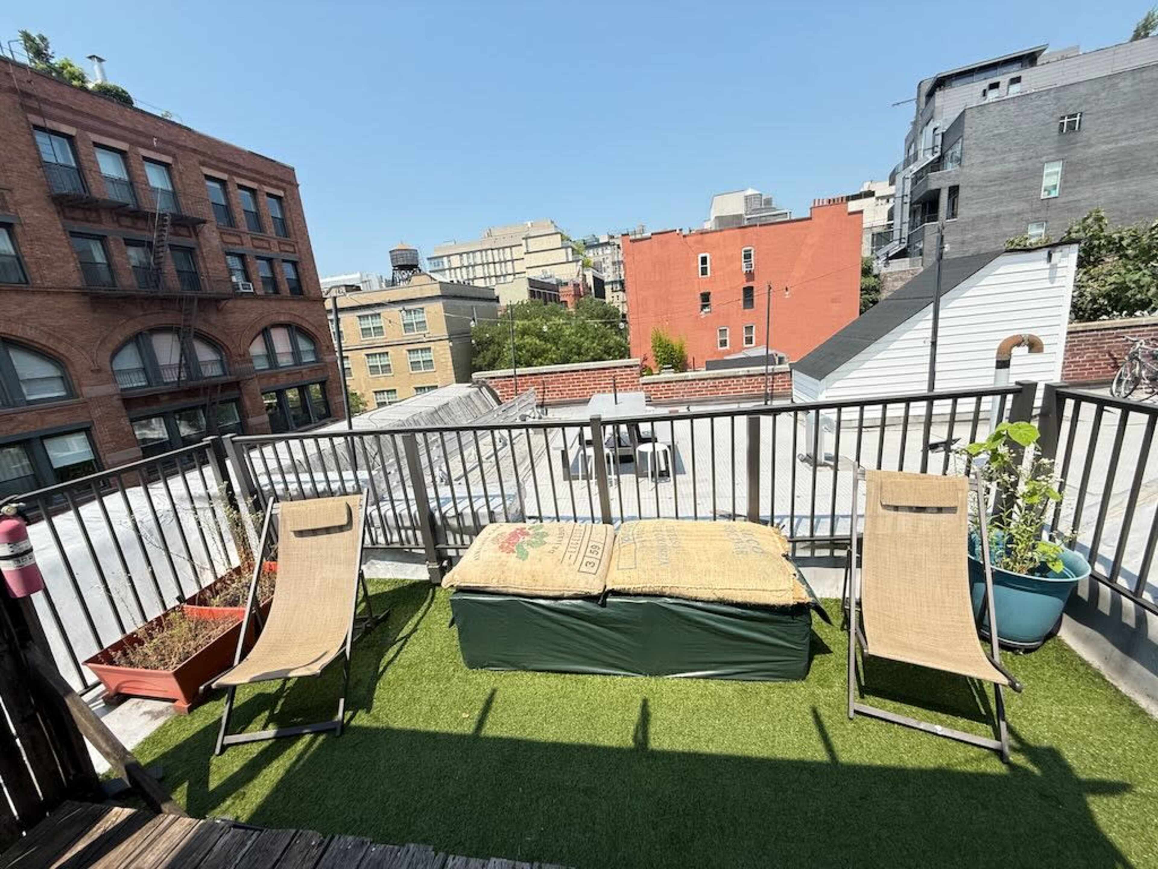 The image shows a rooftop terrace with two lounge chairs, a storage box covered with a tarp, potted plants, and a view of surrounding buildings under a clear blue sky.