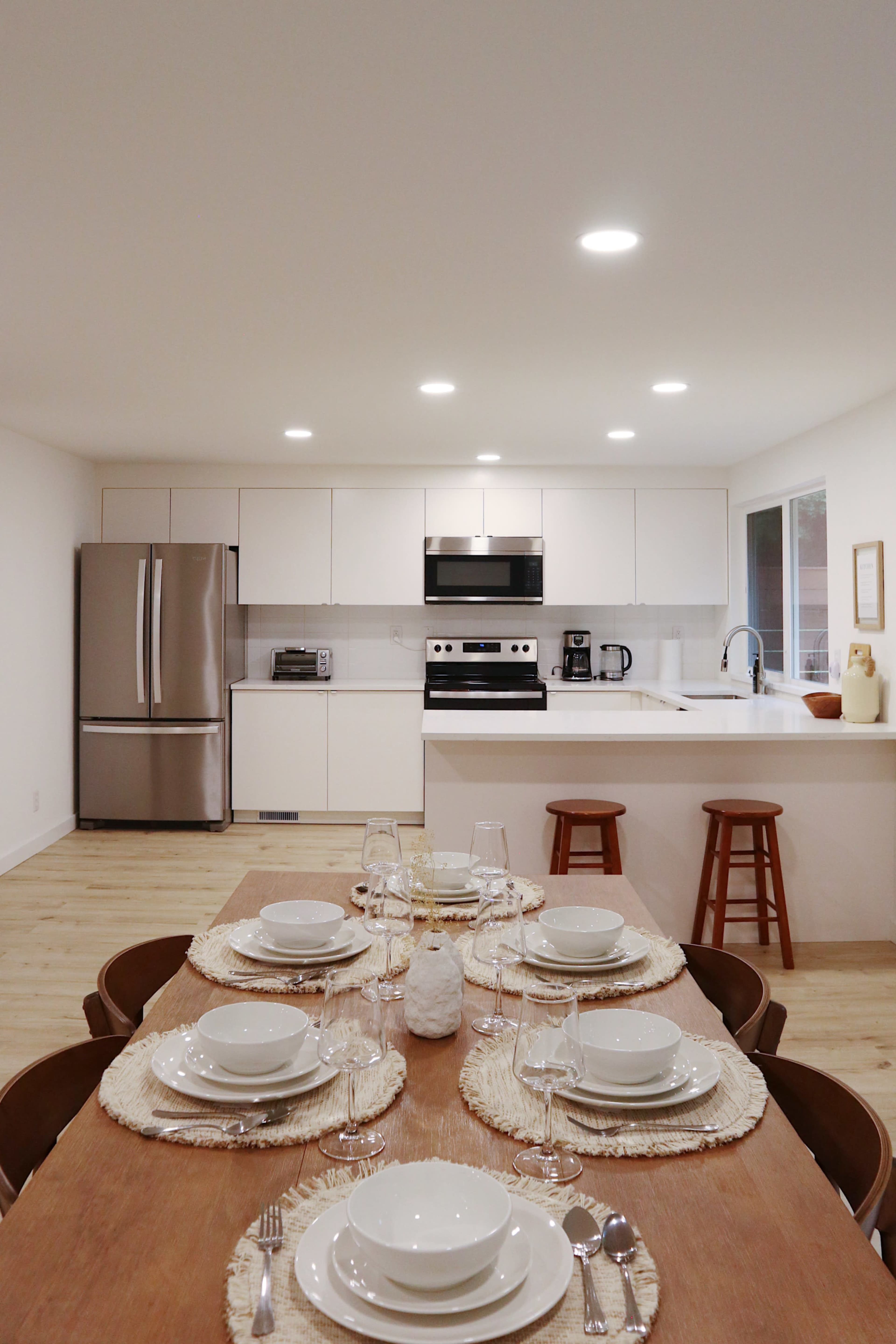A modern kitchen features a dining table set with white plates and glassware, overlooking stainless steel appliances and a breakfast bar.