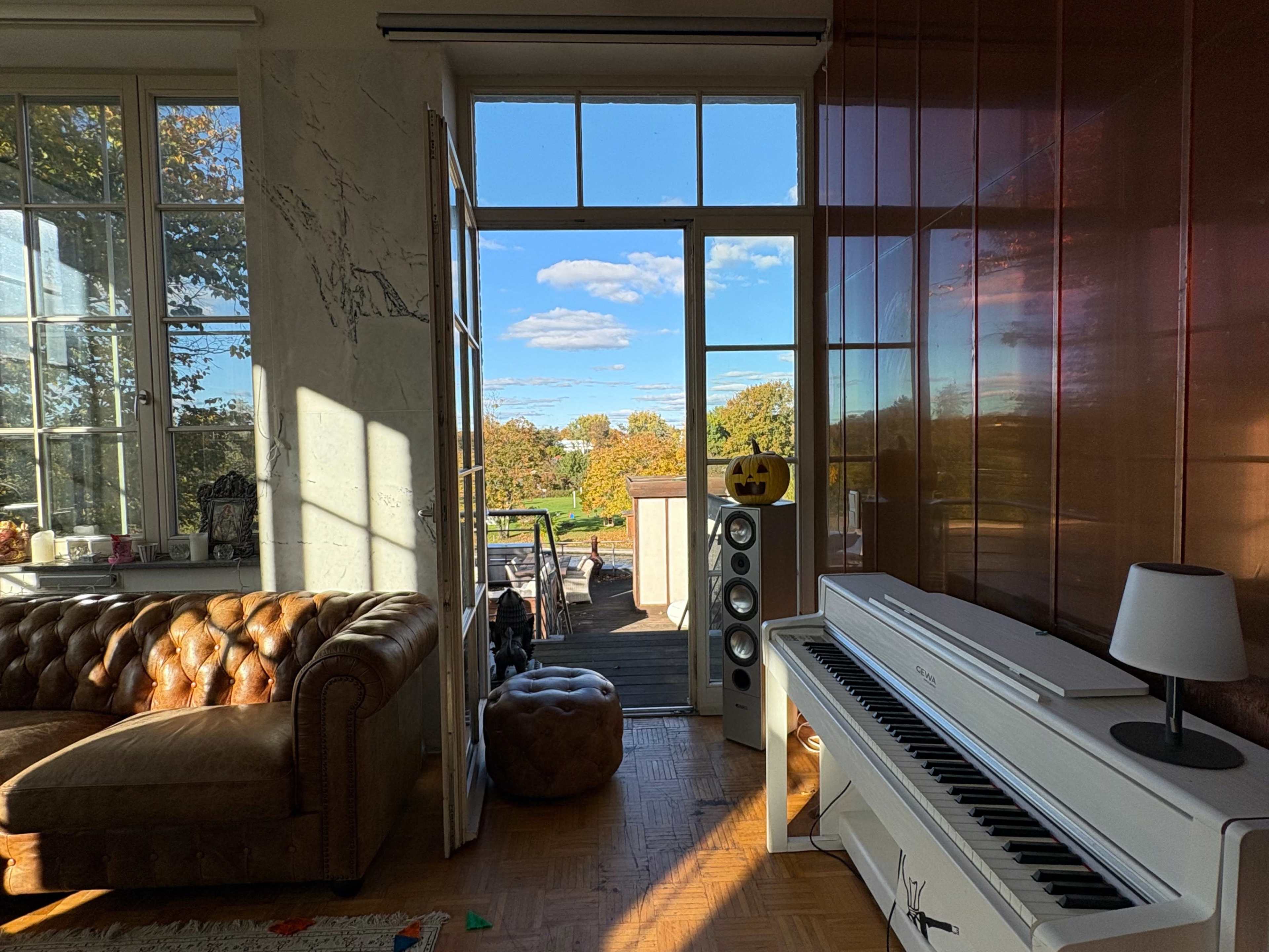 A bright room features a brown leather sofa and a white piano, with large windows showing a view of a sunny outdoor scene.