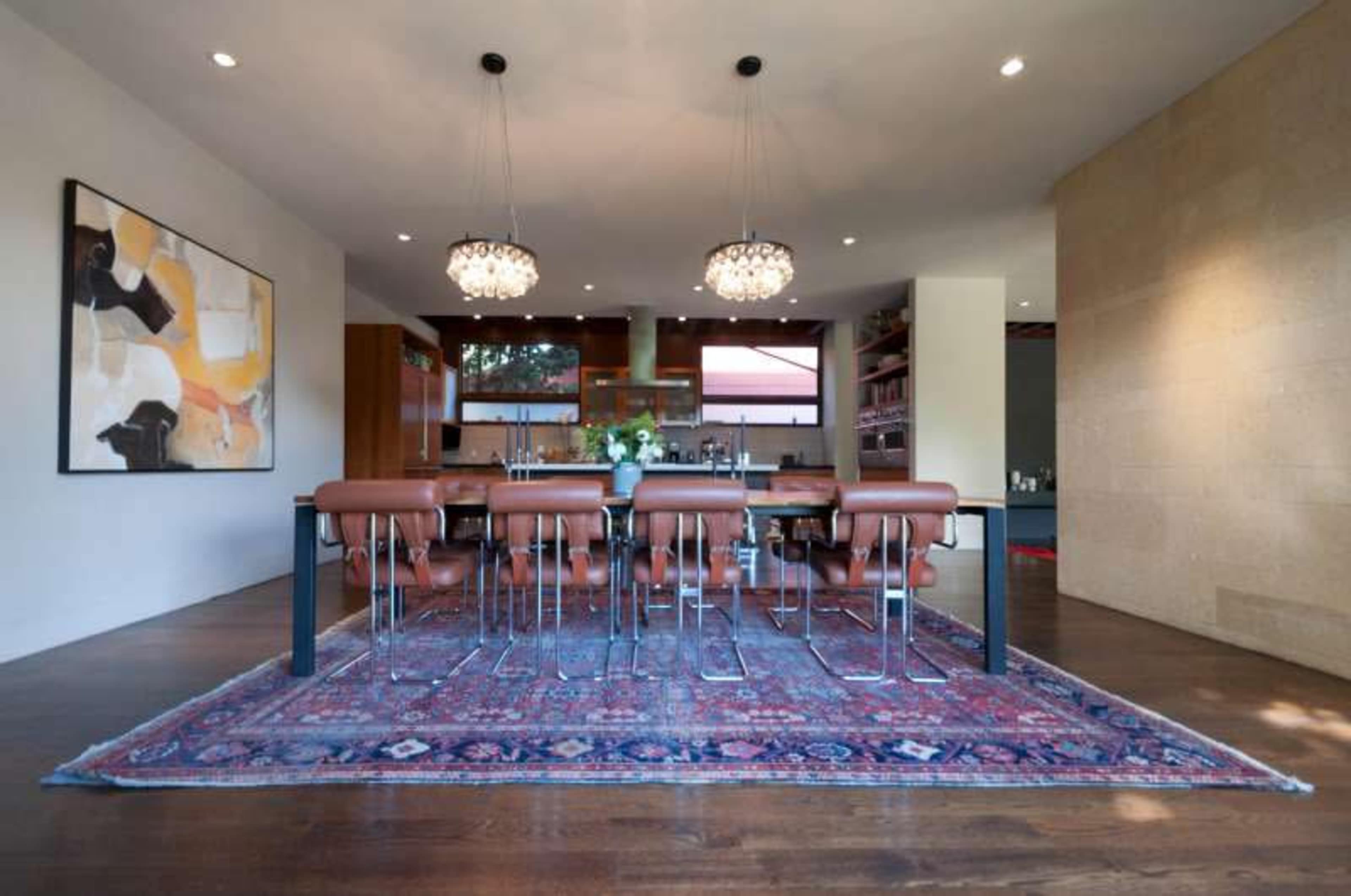 A modern dining area featuring a long table surrounded by brown leather chairs, with a patterned rug on the wooden floor and large pendant lights overhead.