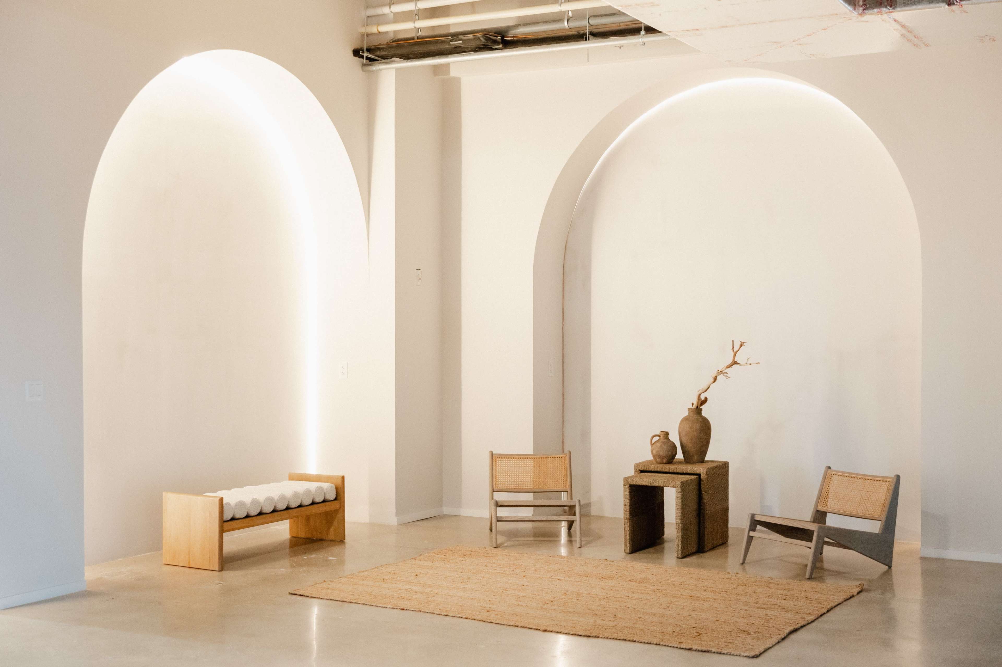 The image shows a minimalist interior with two arched wall alcoves, a wooden bench, two chairs, and a table with a vase and a branch.