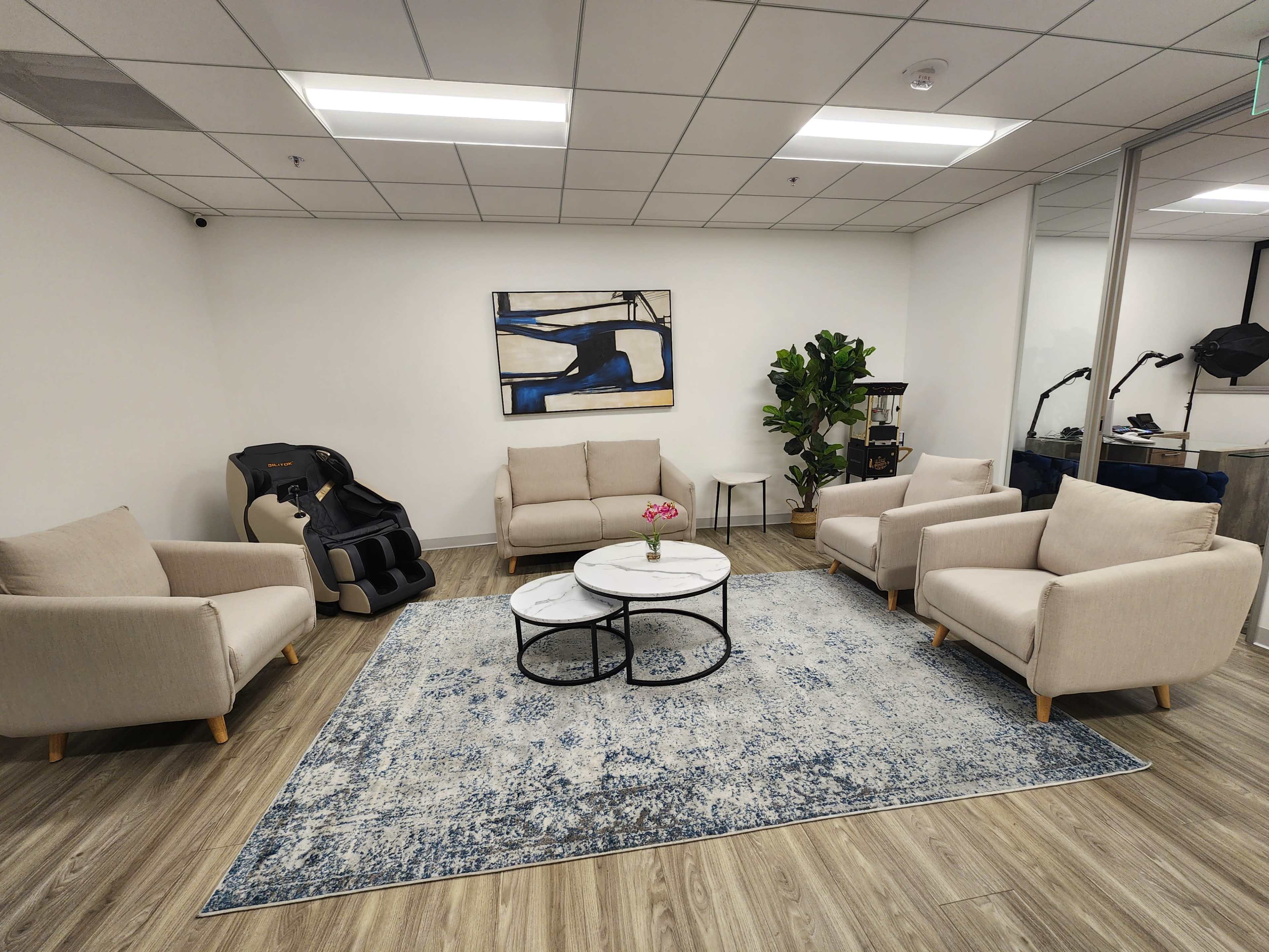 A spacious, modern waiting area with beige sofas, a round coffee table, and a decorative rug, accompanied by a potted plant and wall art.