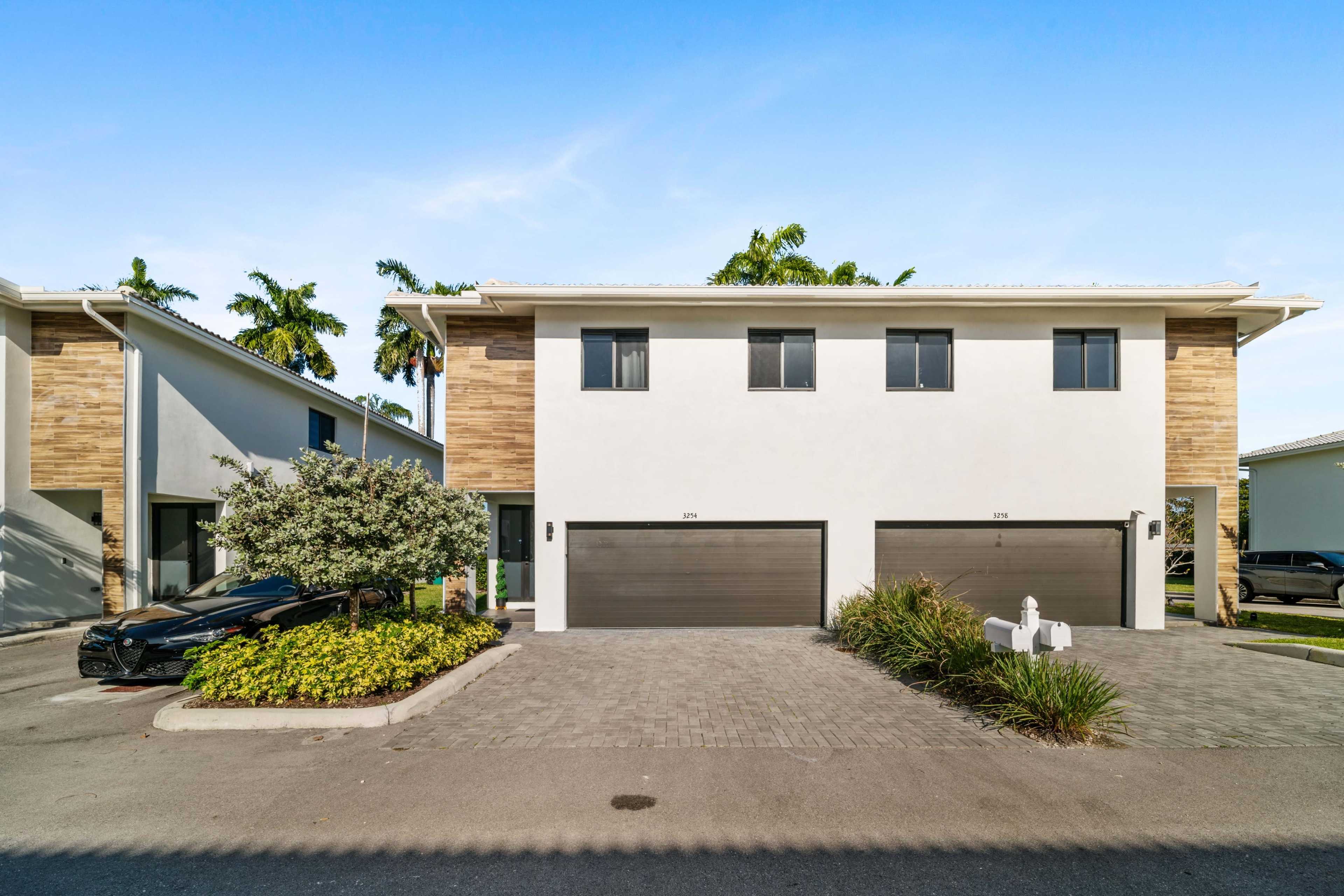 A modern duplex with two separate garages, surrounded by lush landscaping and palm trees.