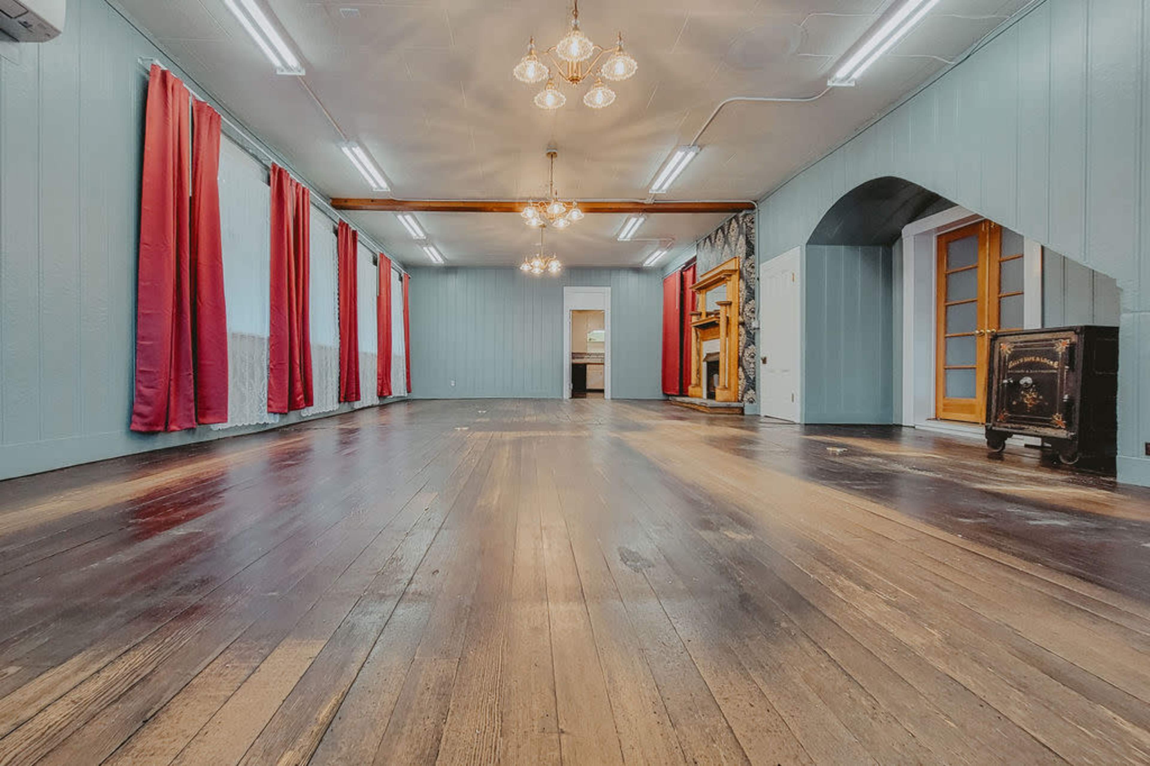 The image shows a spacious room with wooden floors, large windows dressed in red curtains, and several light fixtures hanging from the ceiling.
