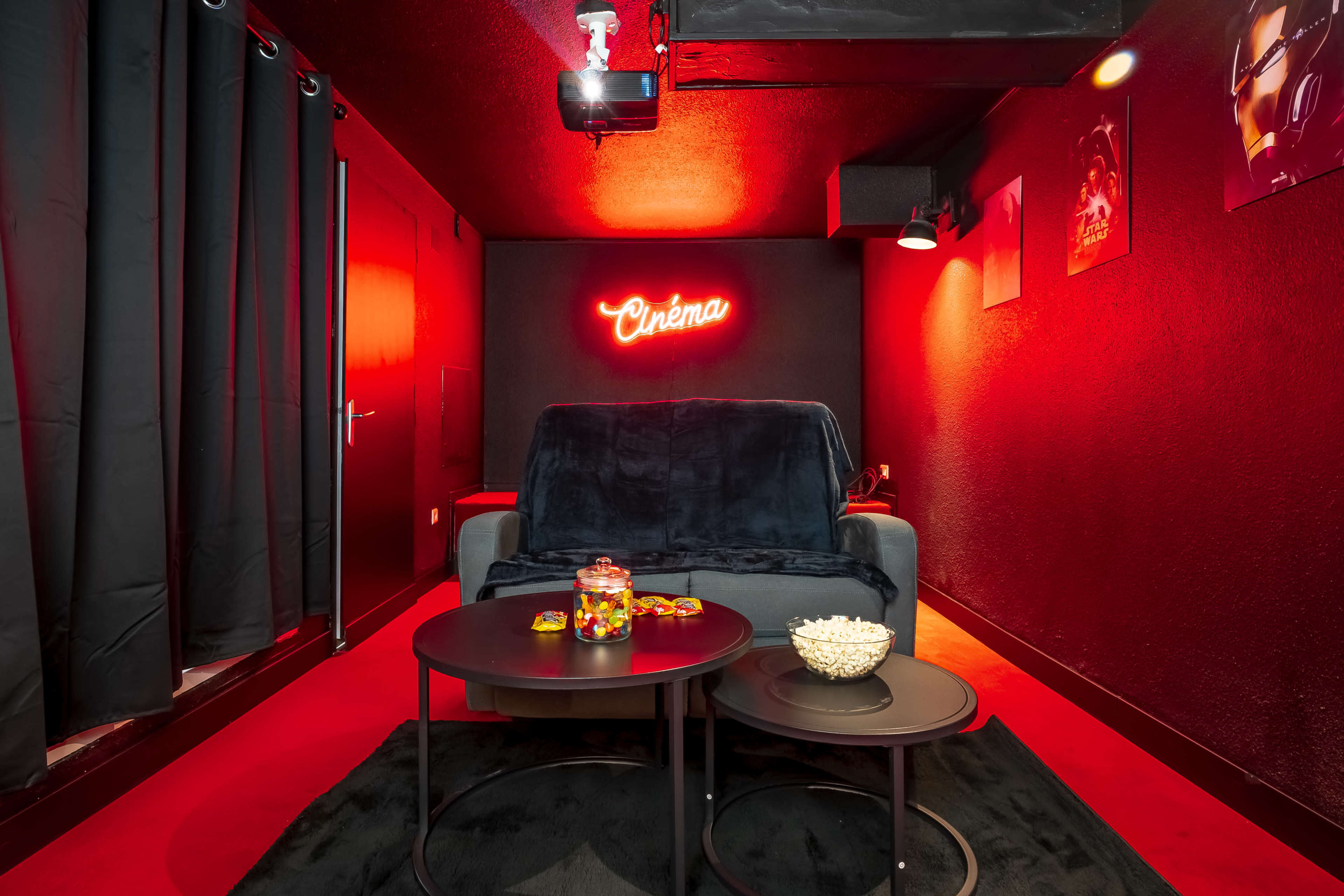 A small home cinema room with a gray couch, black curtains, and red walls, featuring a neon "Cinema" sign and two tables with snacks.