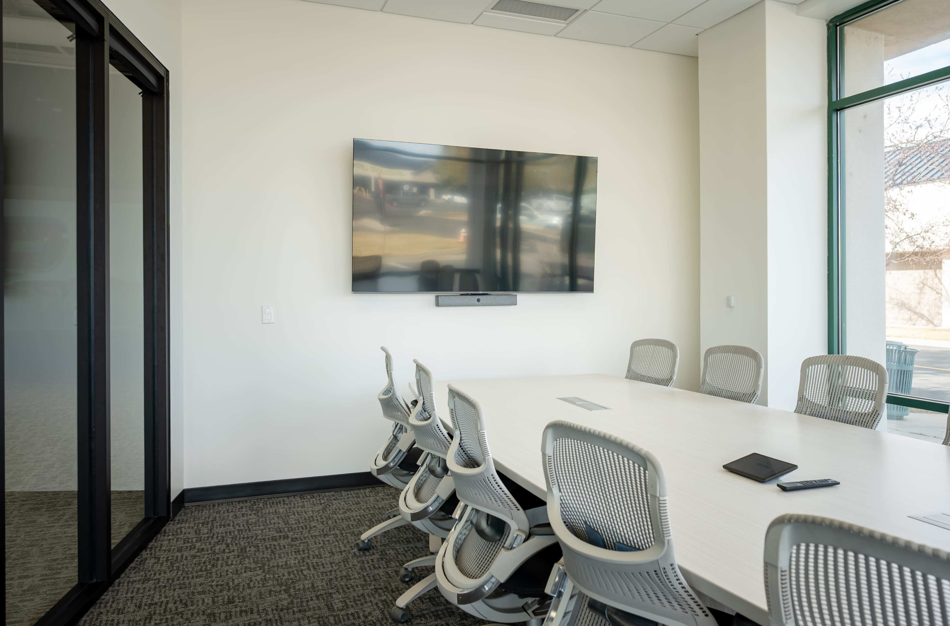 The image shows a modern conference room with a large wall-mounted TV, a long white table, and several ergonomic chairs.