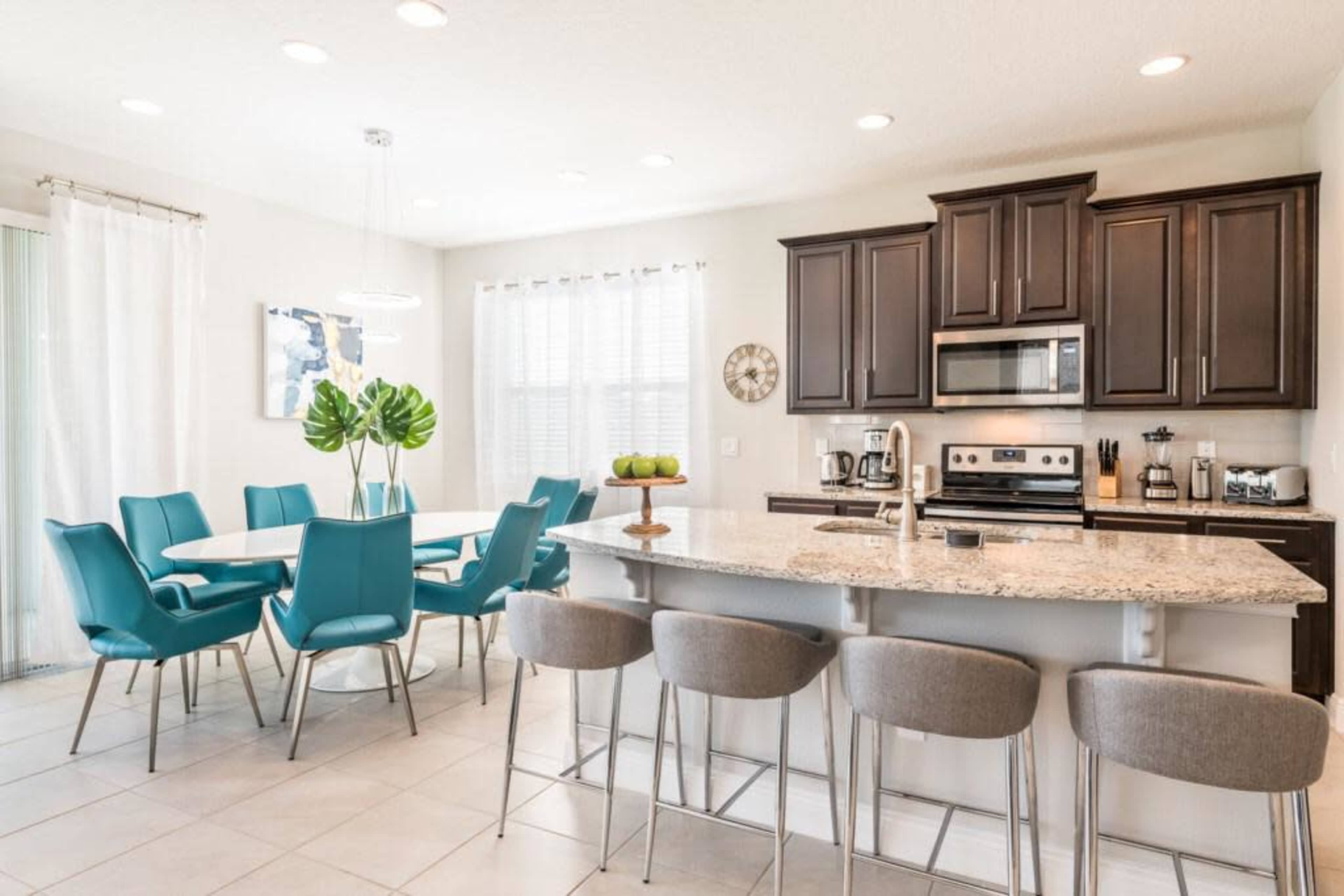 A modern kitchen features dark cabinets, a granite island with gray bar stools, and a dining area with a large table and teal chairs.