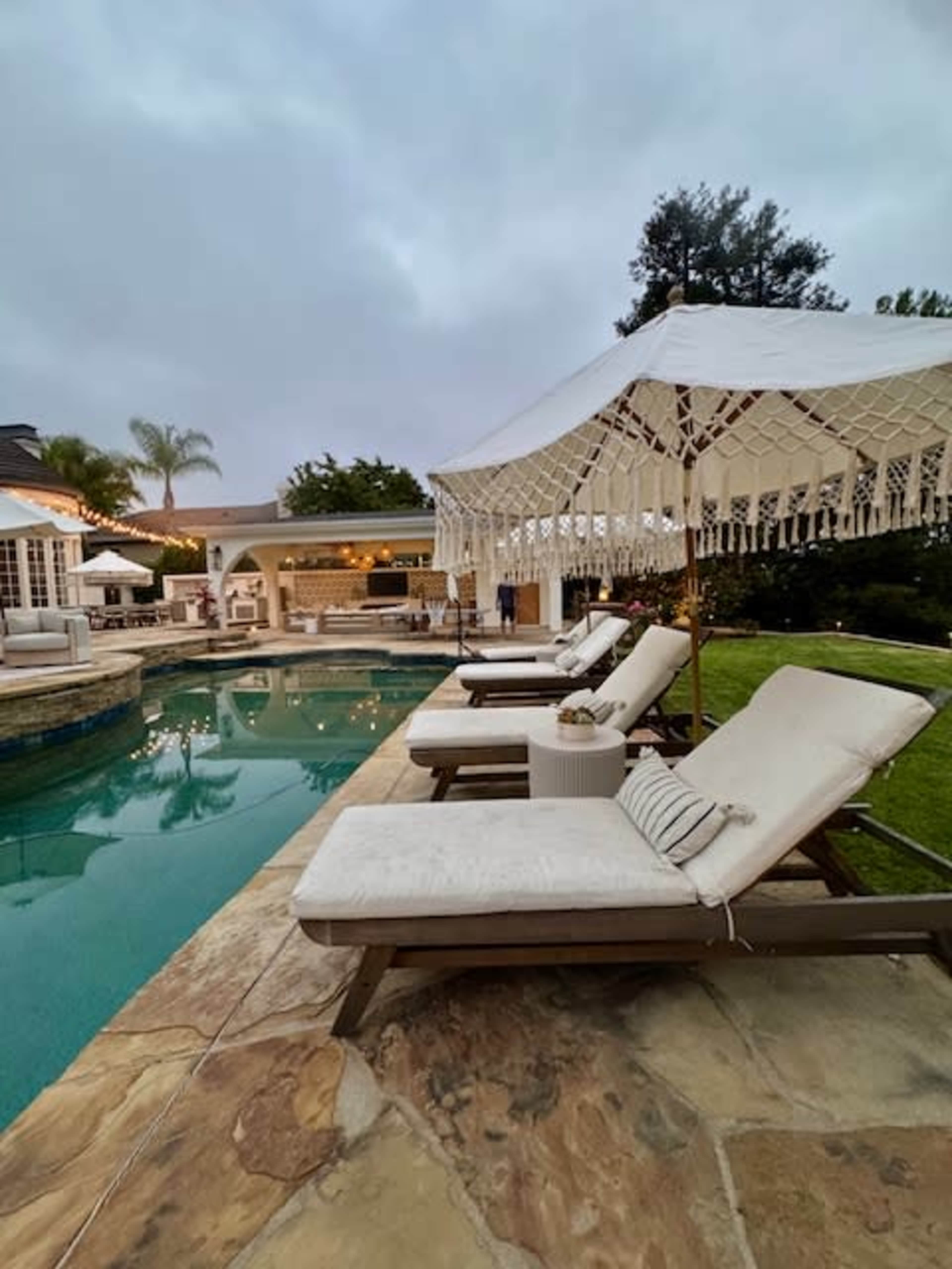 A set of lounge chairs are arranged near a poolside area with a tented structure in the background, surrounded by greenery and a cloudy sky.