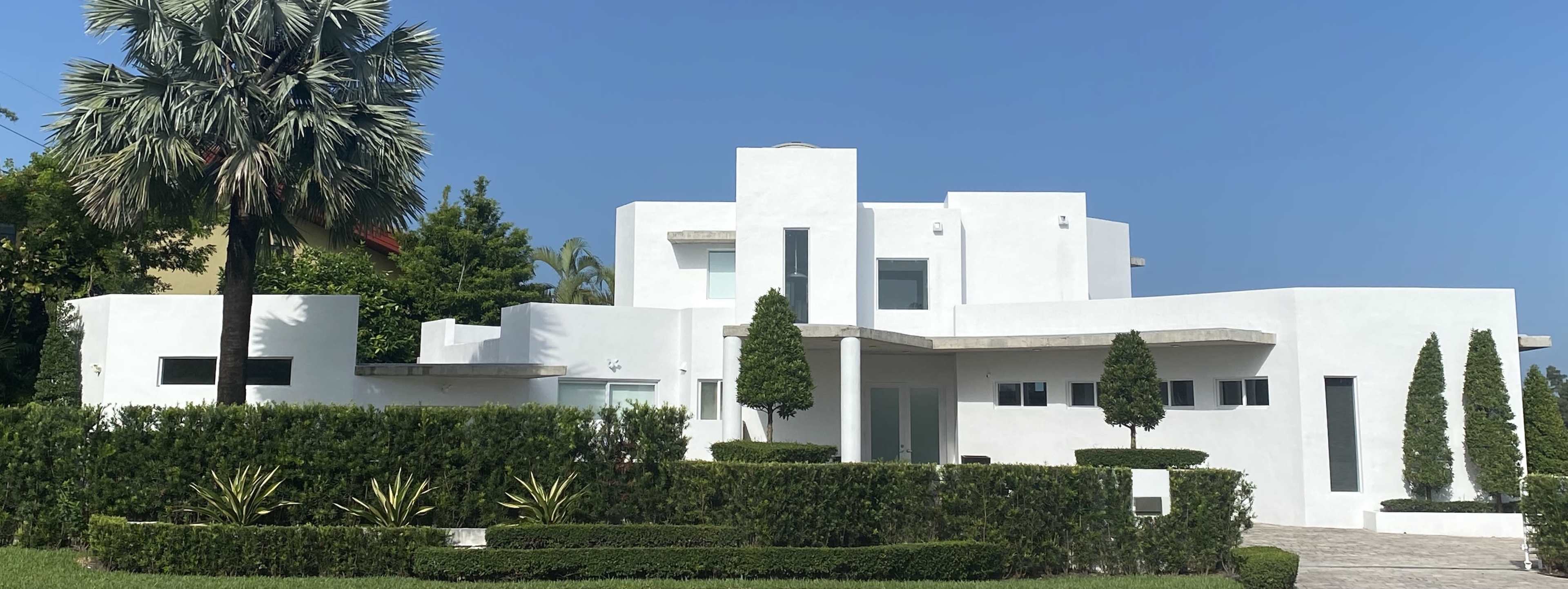 The image shows a modern, white stucco house with geometric shapes and landscaped greenery.