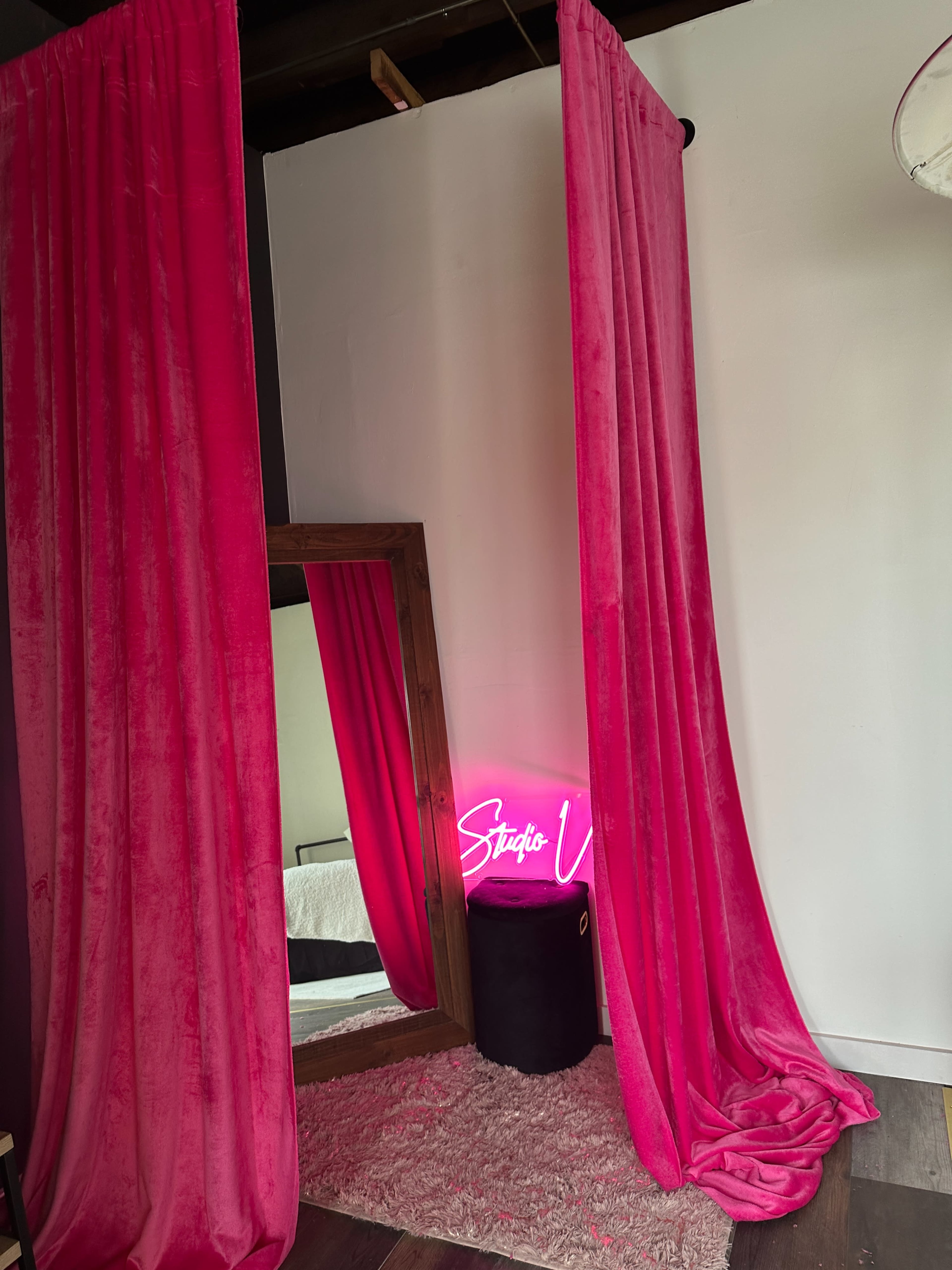 The image shows a corner of a room with pink velvet curtains, a large mirror, a neon sign reading "Studio V," and a black stool on a textured rug.