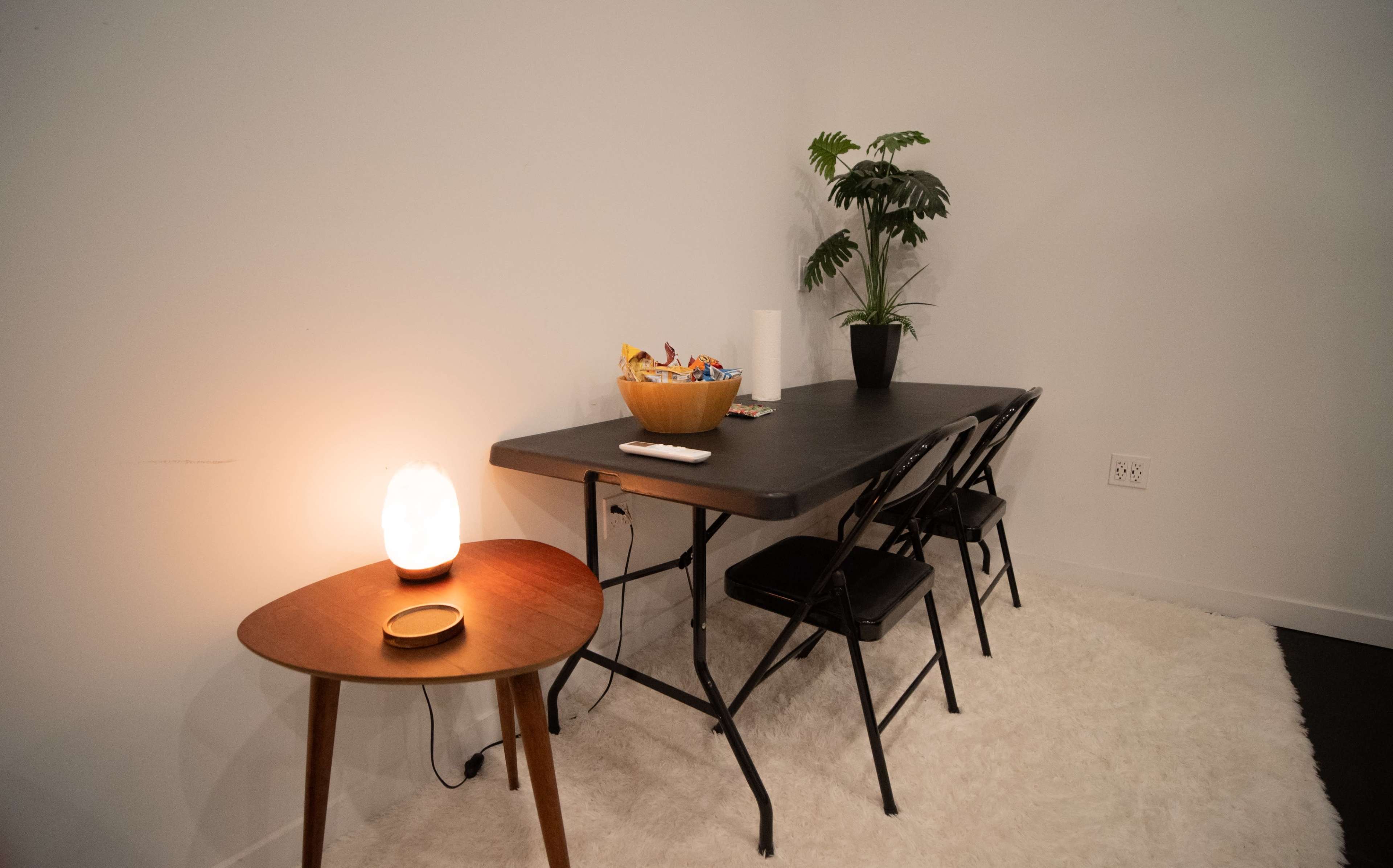 A simple dining area features a dark folding table with three chairs, a bowl of snacks, a potted plant, and a small side table with a lamp.