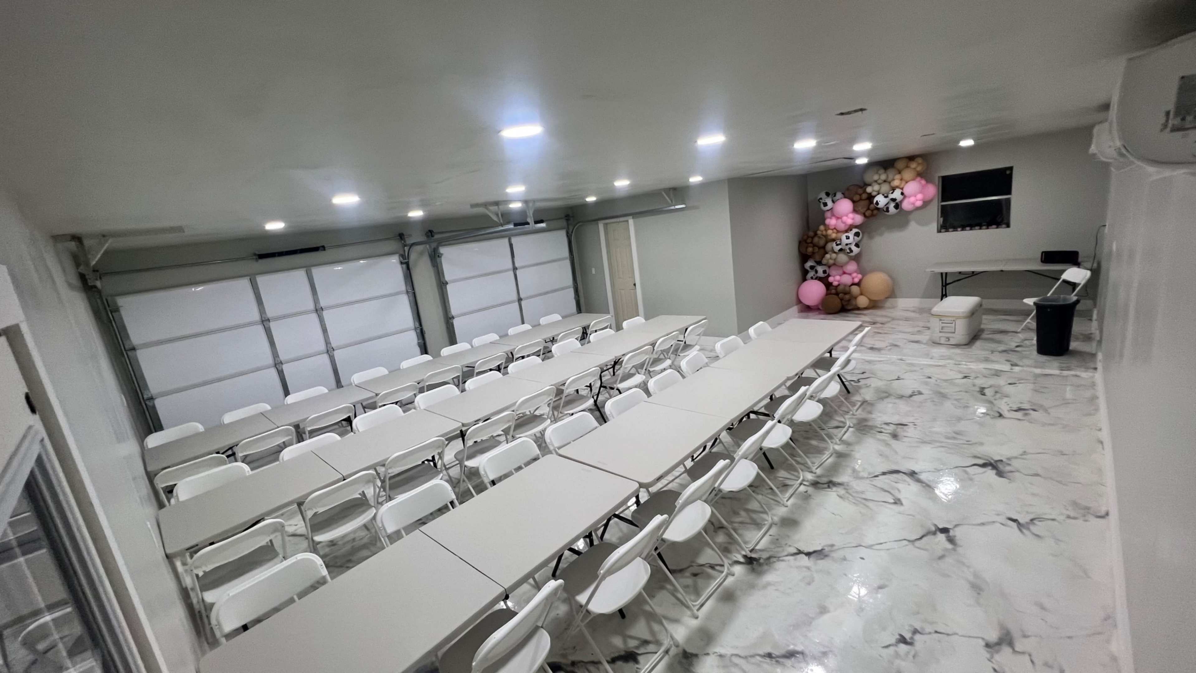 A spacious rental room with multiple rows of tables and chairs, a decorative wall of balloons, and large garage doors in the background.