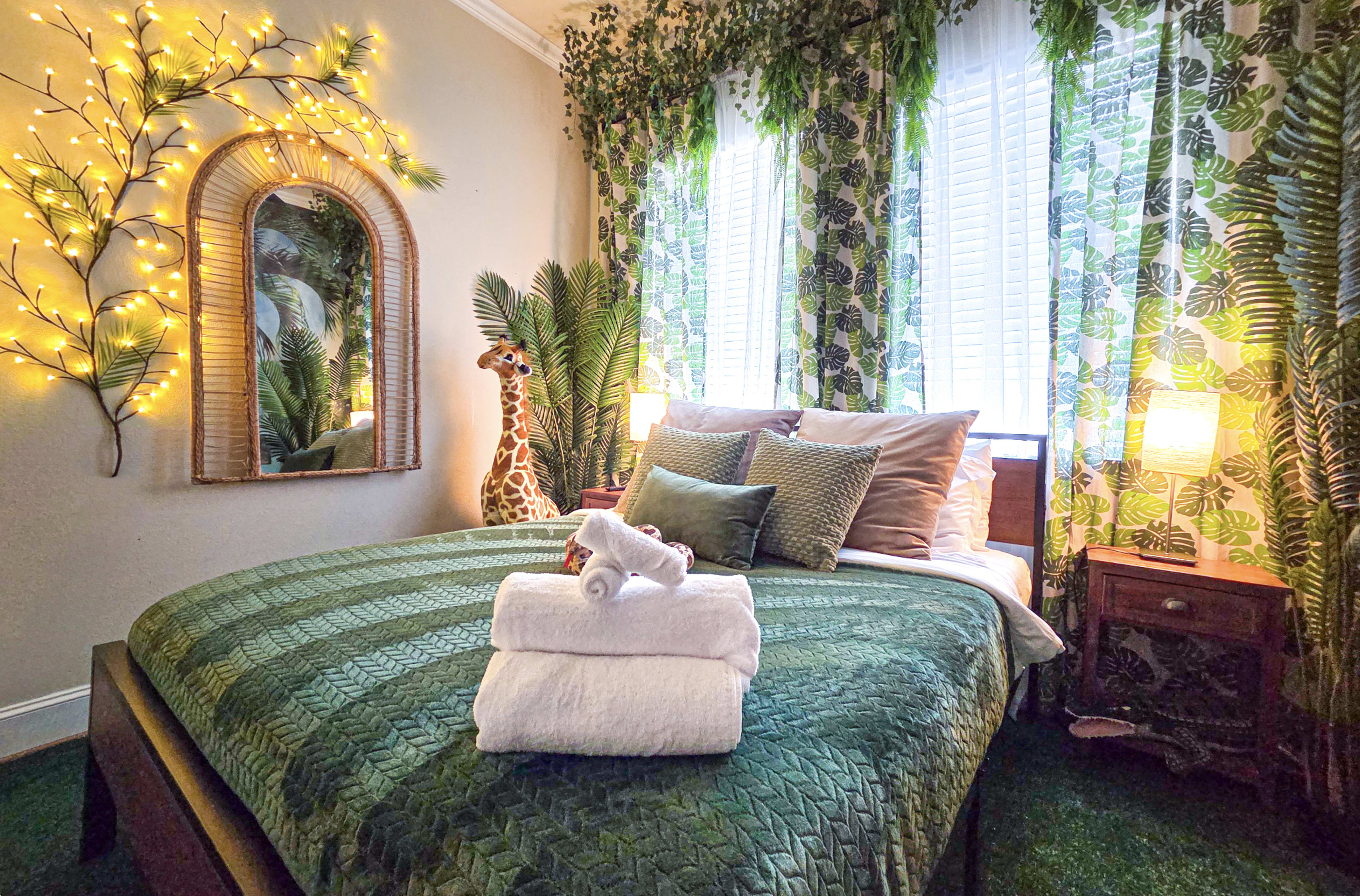 The image shows a cozy bedroom decorated with green bedding, a giraffe statue, and plants, illuminated by warm fairy lights and natural light from the windows.